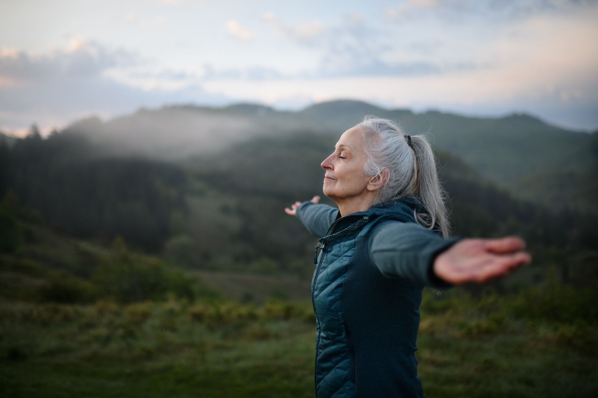 Woman in a teal jacket stands with arms outstretched in a misty mountain landscape at dawn