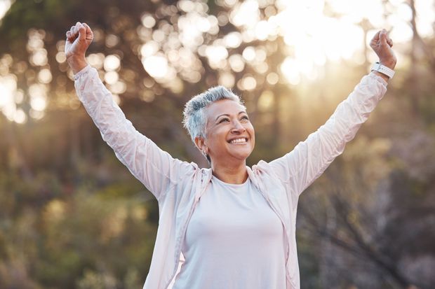 A smiling person with short gray hair stands outdoors with arms raised in a gesture of celebration at sunset.