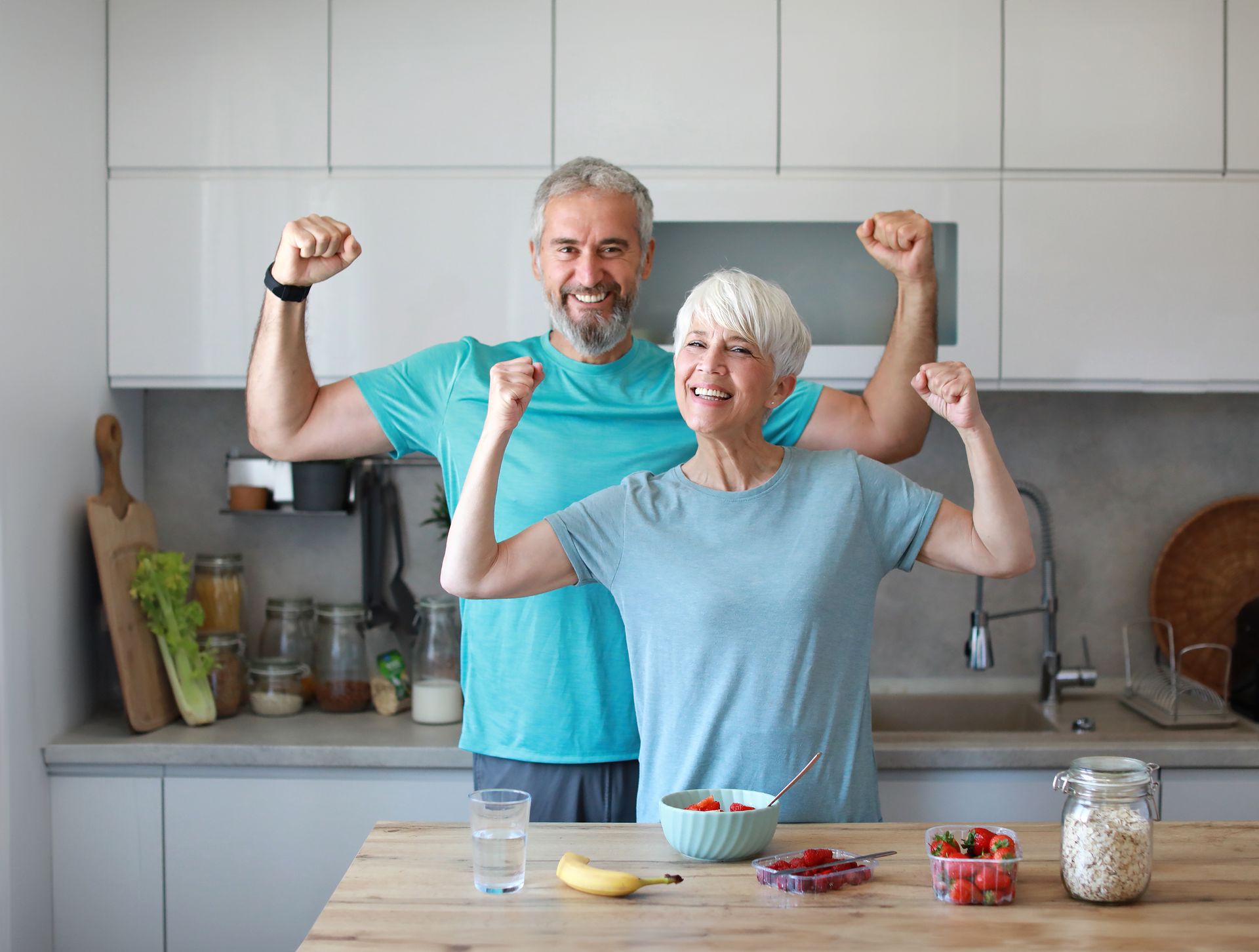 Two people flexing in a bright kitchen, smiling behind a counter with breakfast items.