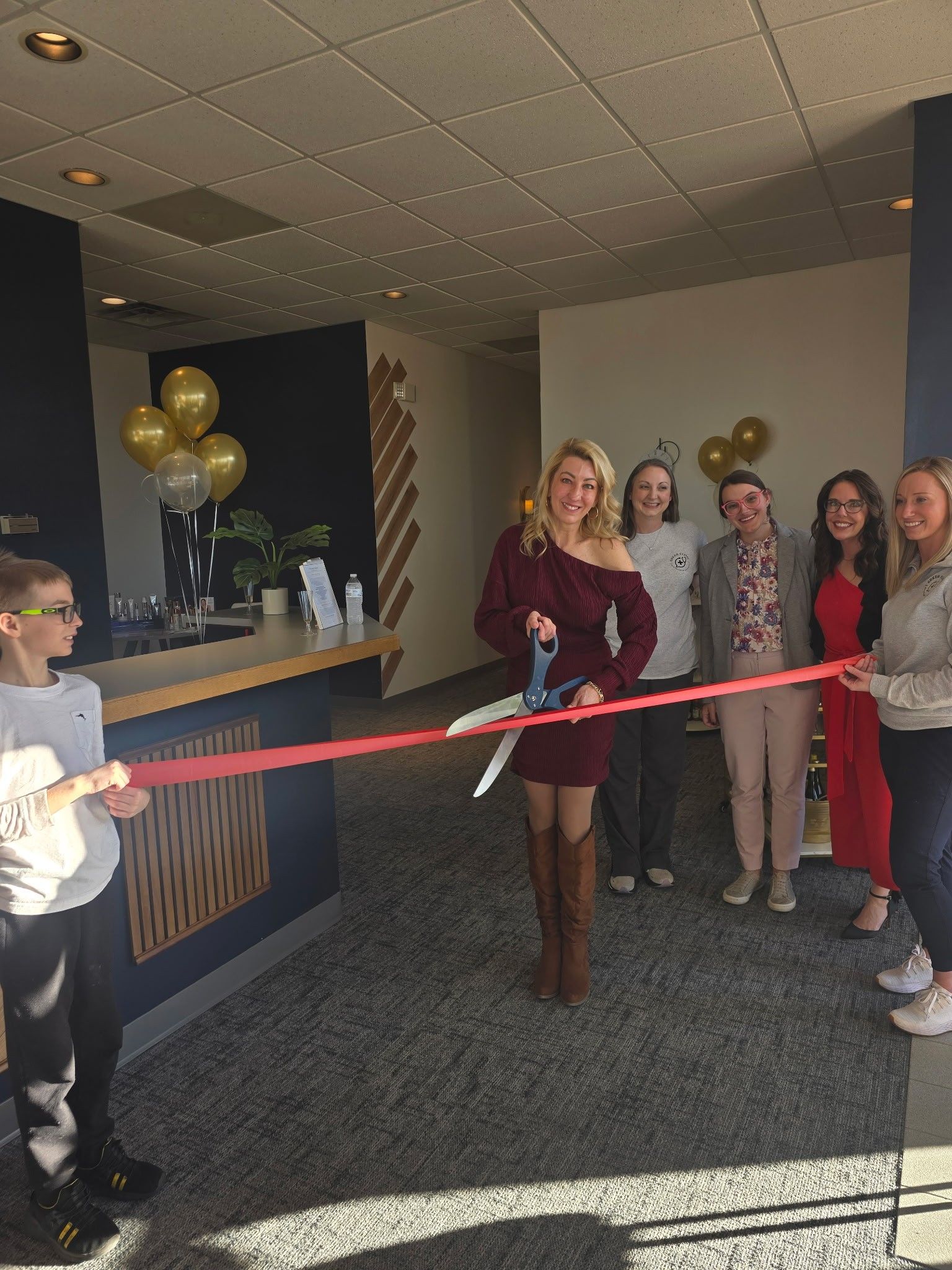 People cutting a ribbon at a business opening. Blond woman cuts ribbon with large scissors, others watch. Balloons and a bar visible.