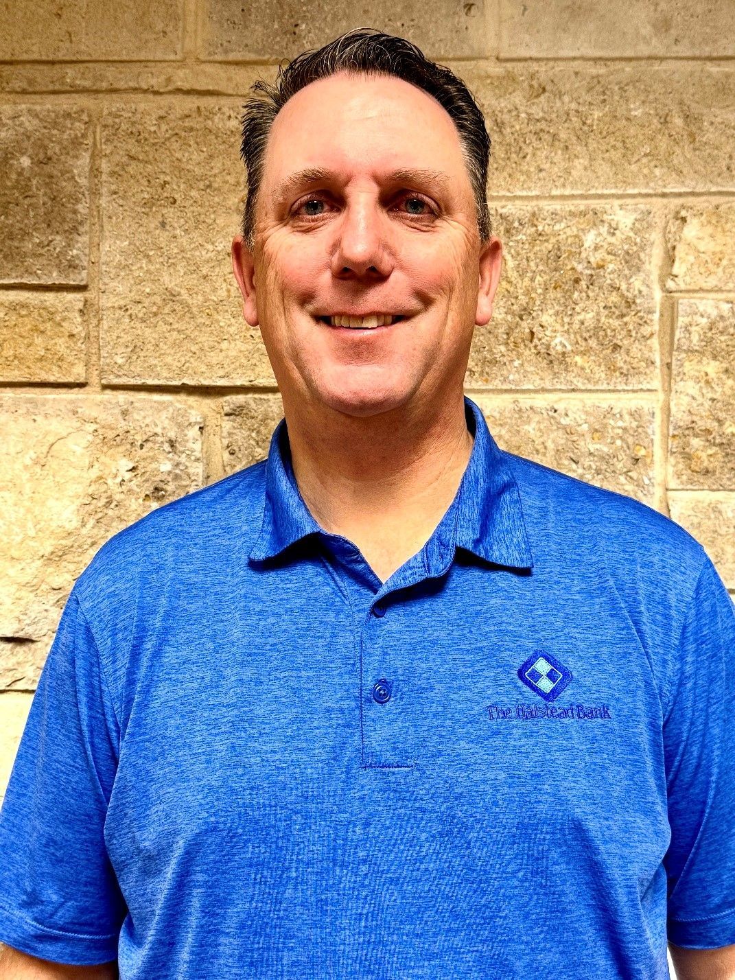 Man in blue polo shirt smiles, standing in front of a tan brick wall.
