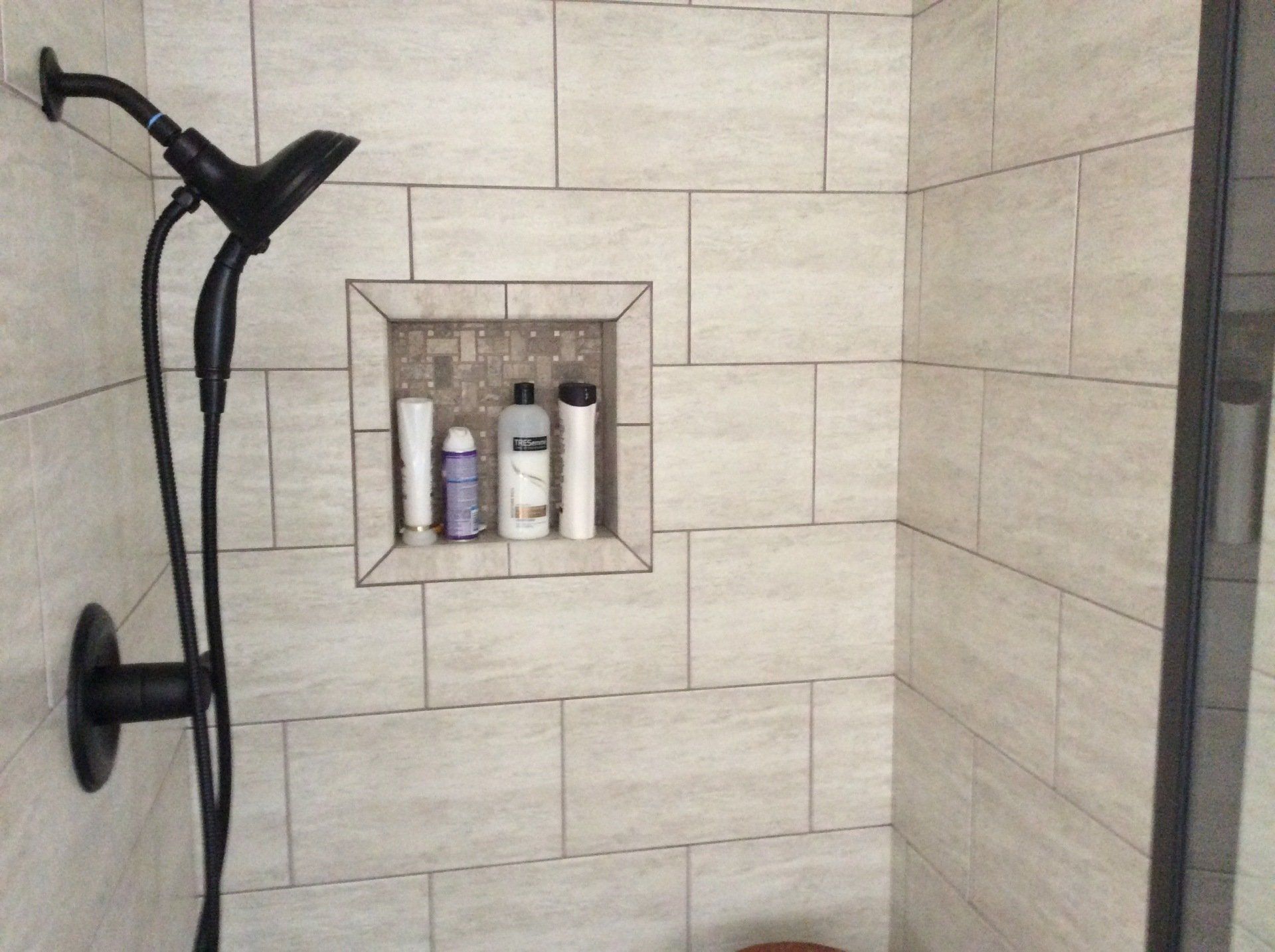 A shower with a black shower head and a shelf with bottles in it