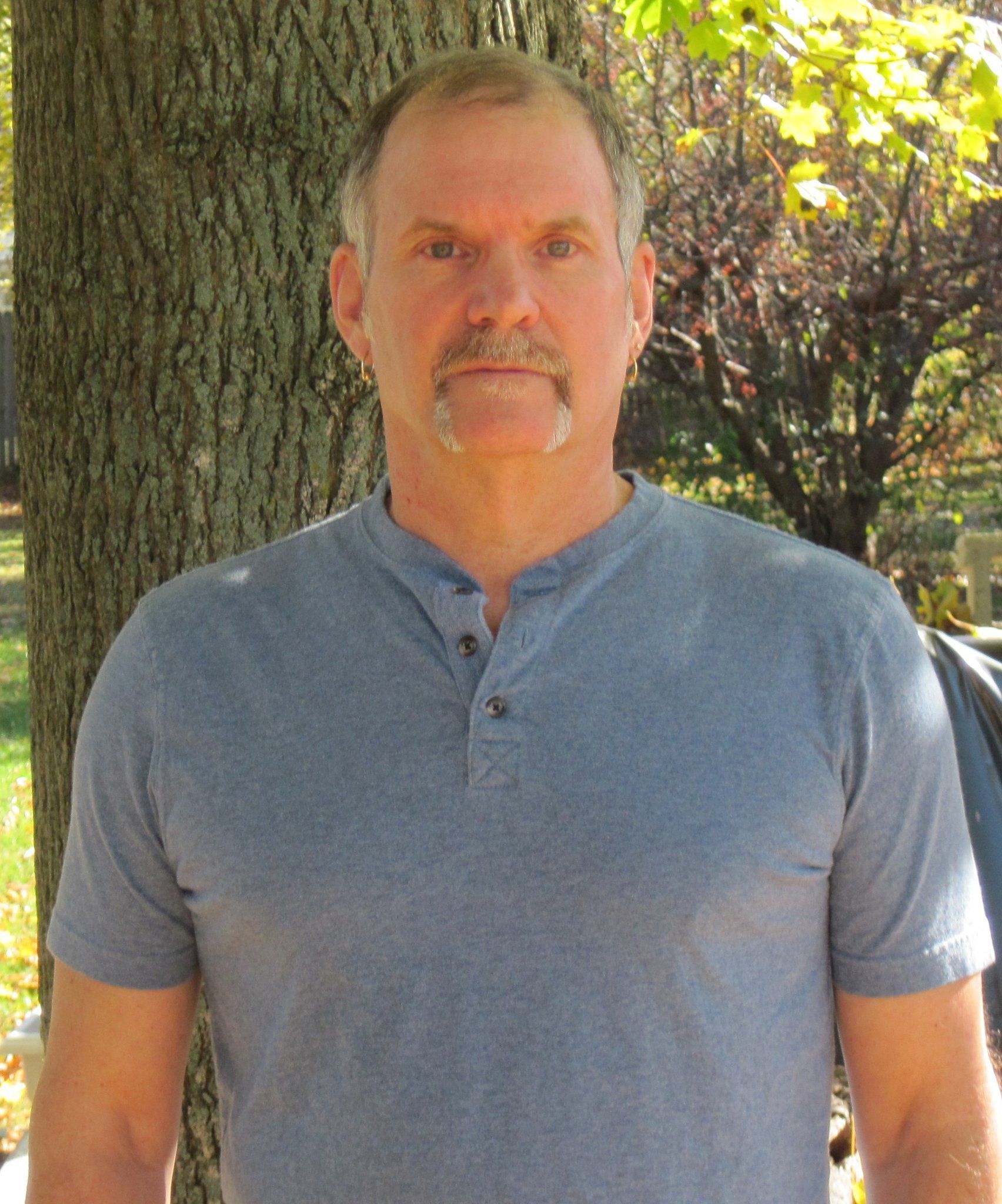 A man with a beard wearing a grey shirt is standing in front of a tree
