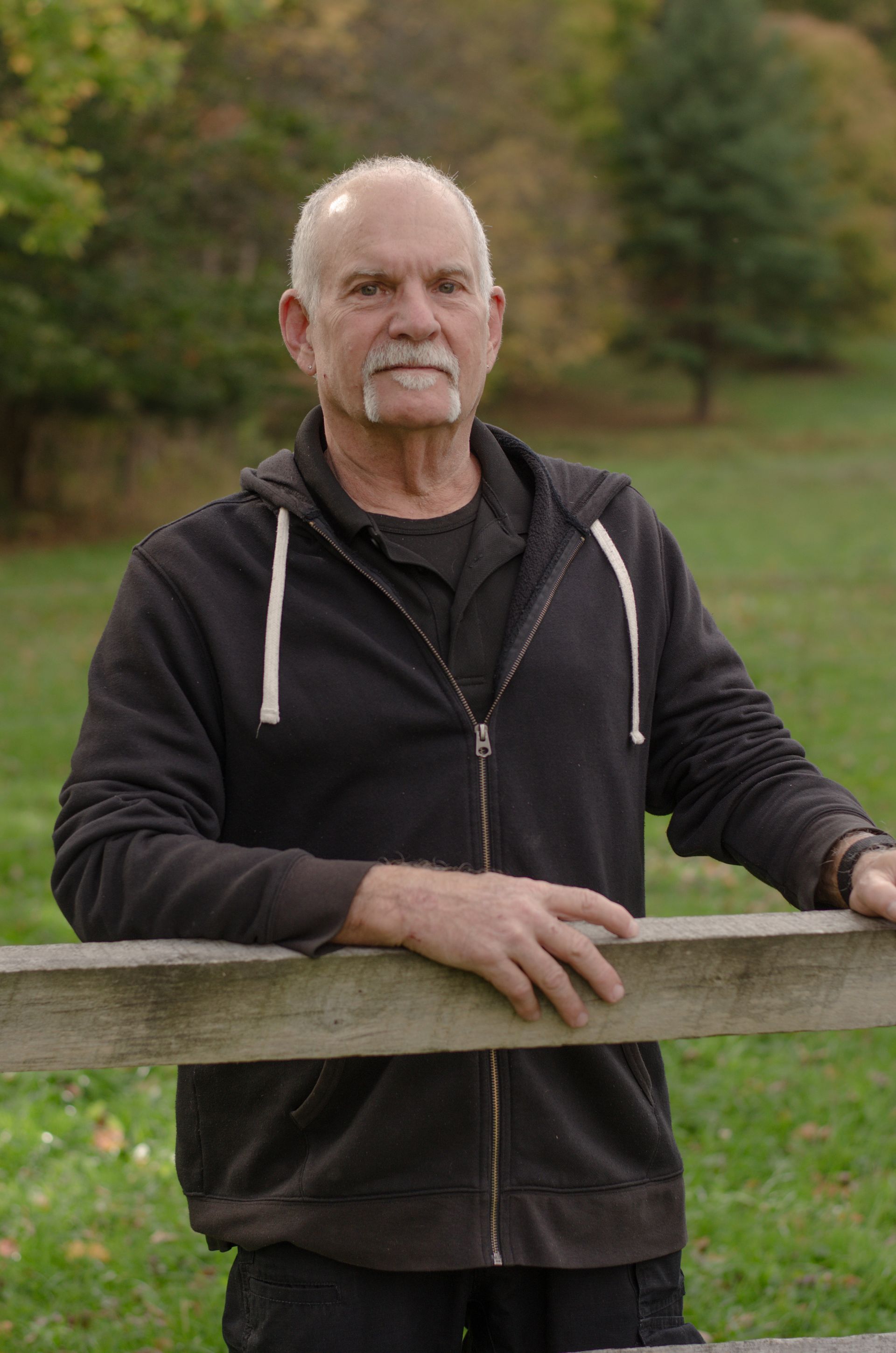 A man with a beard wearing a grey shirt is standing in front of a tree