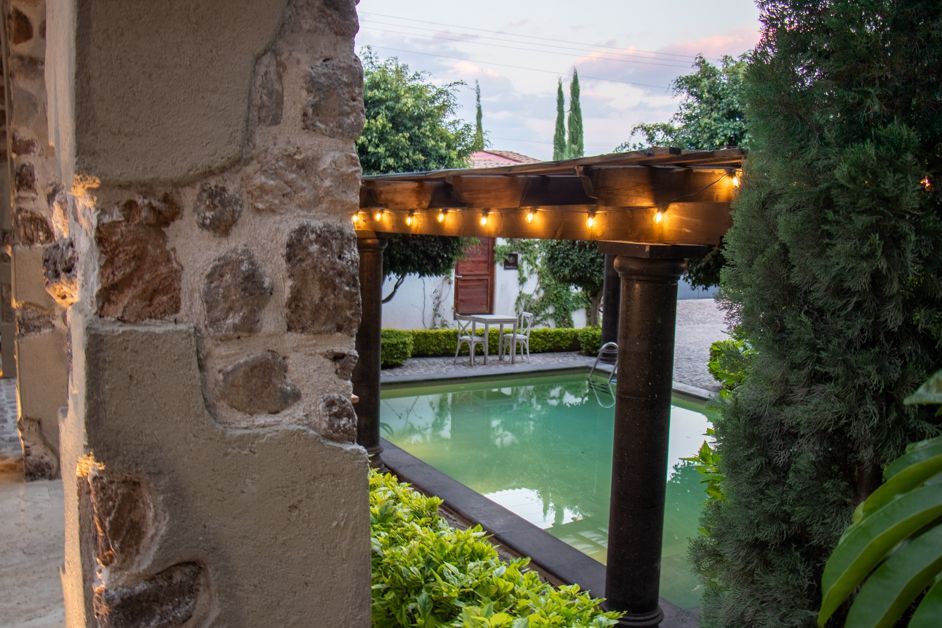 Stone wall, pool with wooden pergola lit by string lights; outdoor setting.