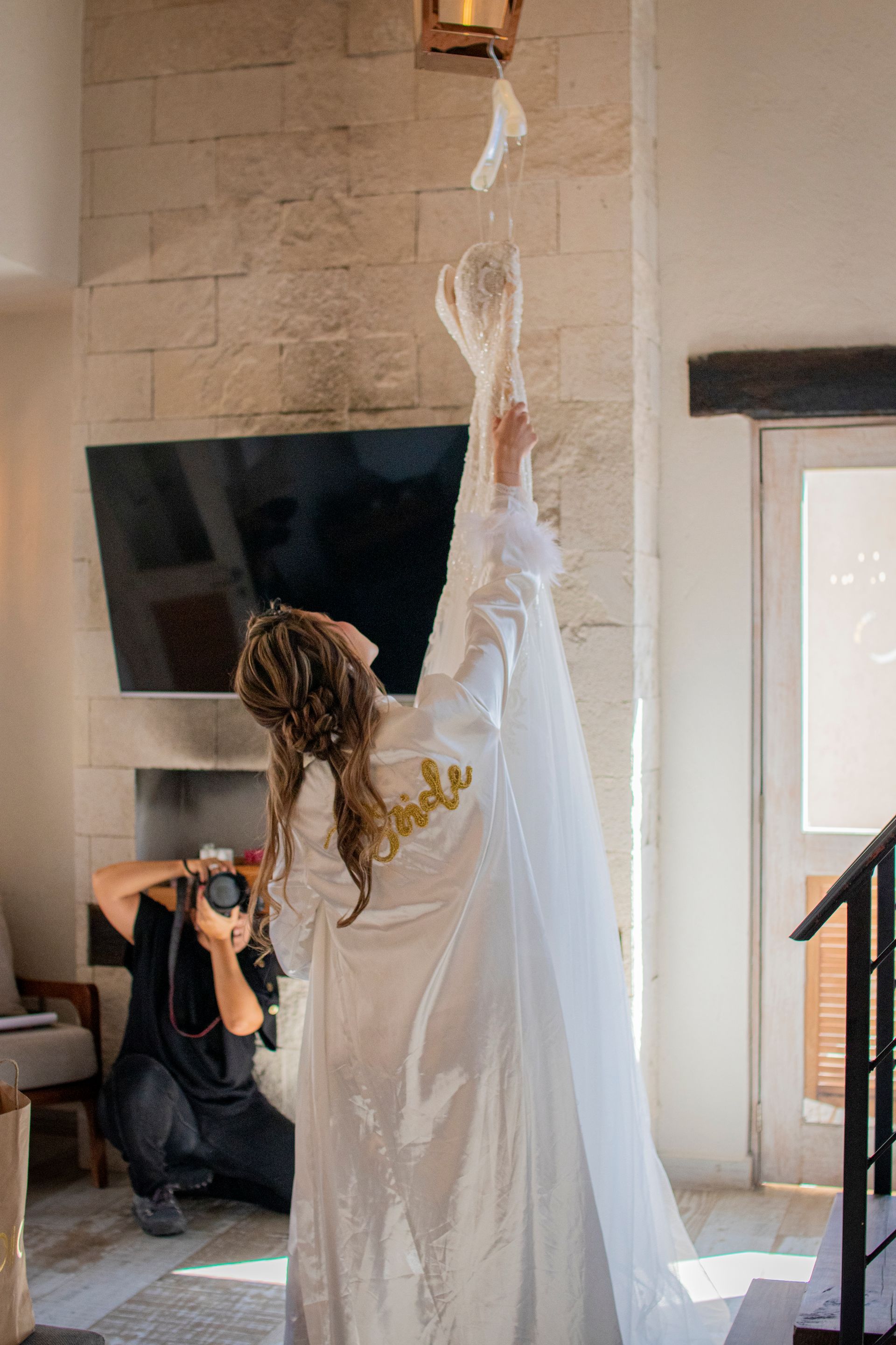 Woman tossing her wedding dress up in air, looking up. Photographer taking photo. Inside room with stone wall.
