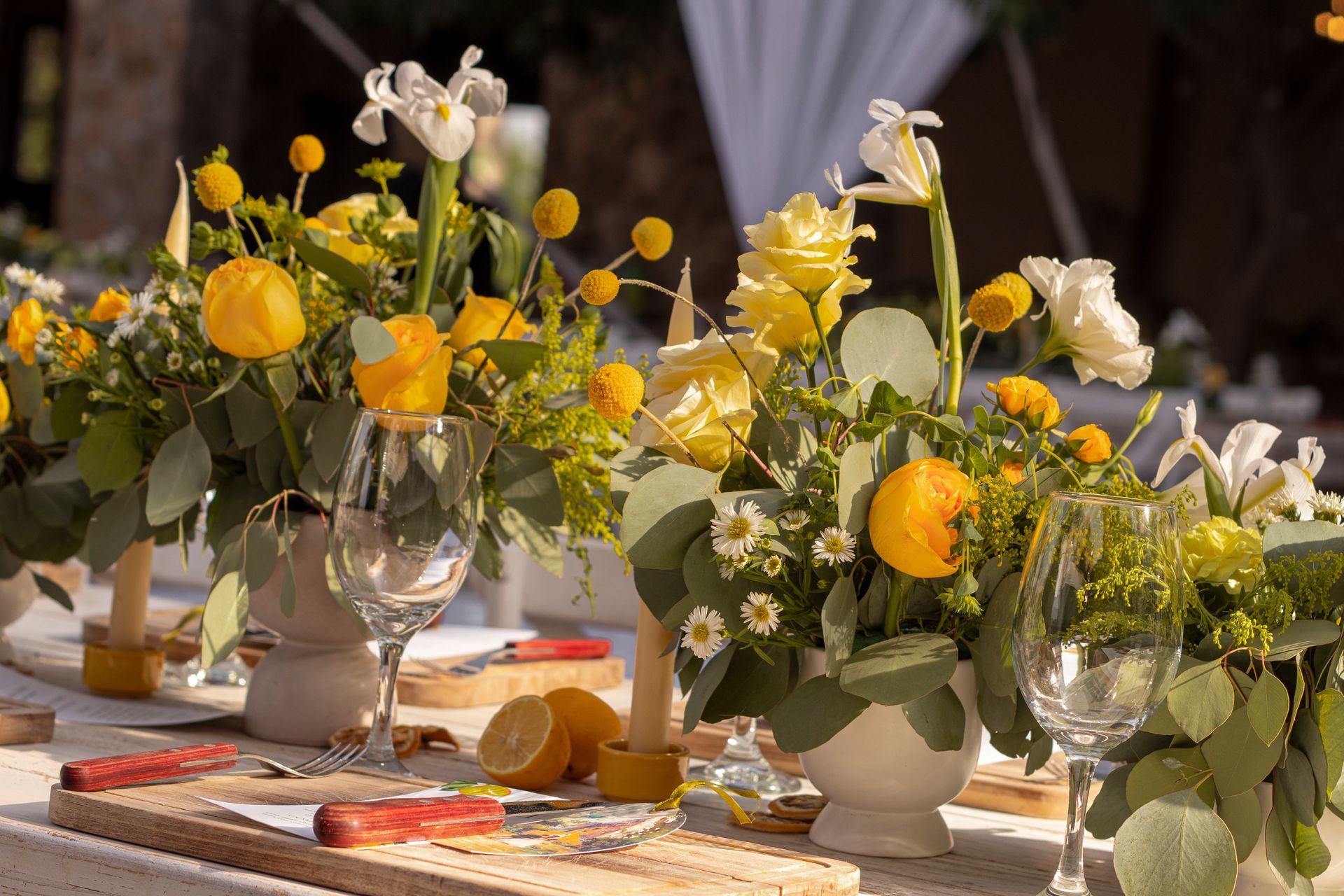 Centerpieces of yellow roses, lemons, and white flowers on a table set for a meal.