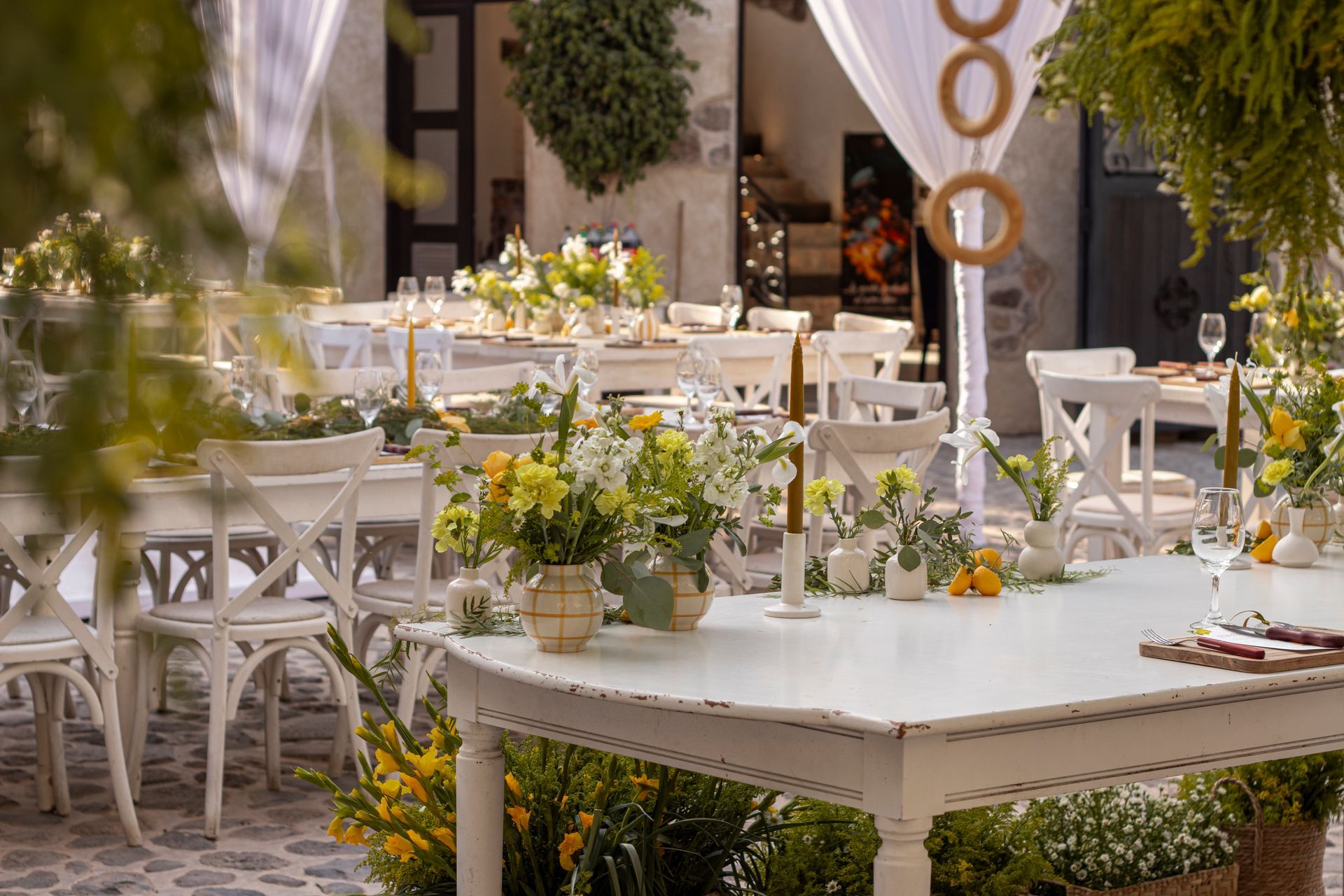 White tables set for a gathering, decorated with flowers and candles in an outdoor courtyard.