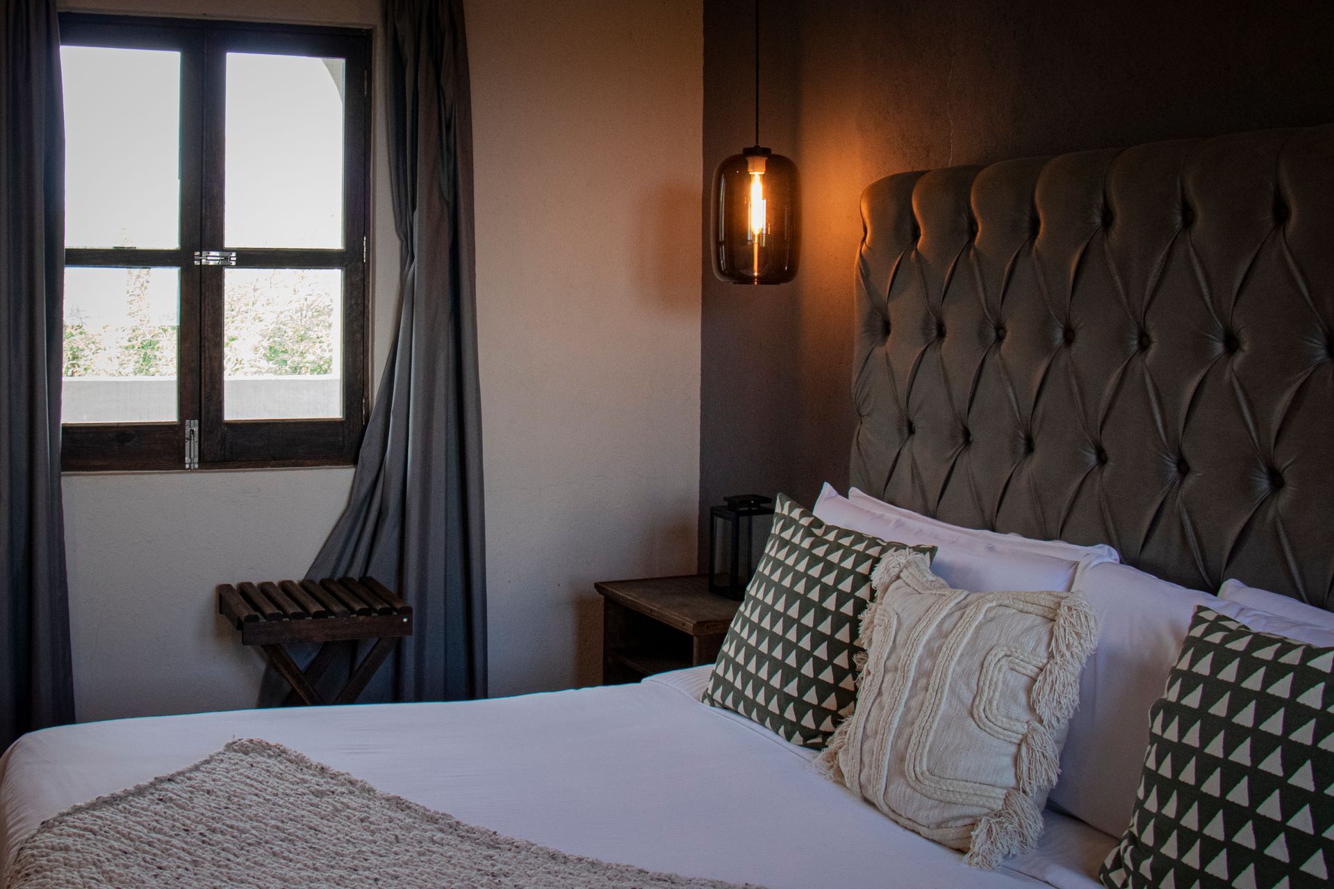 Bedroom with tufted headboard, hanging light, window, and patterned pillows.