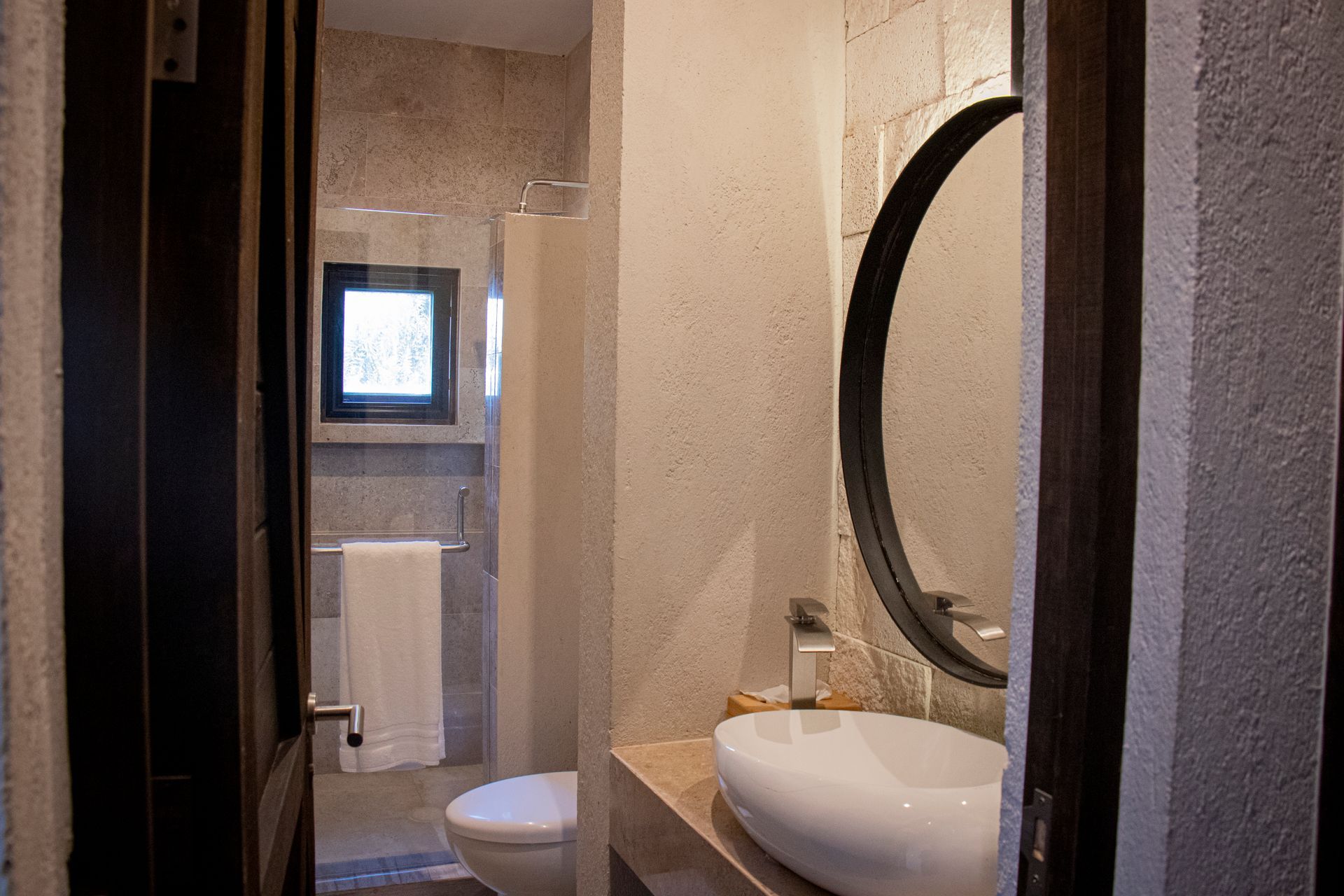 Bathroom interior with a round mirror, vessel sink, toilet, and open door to a shower.