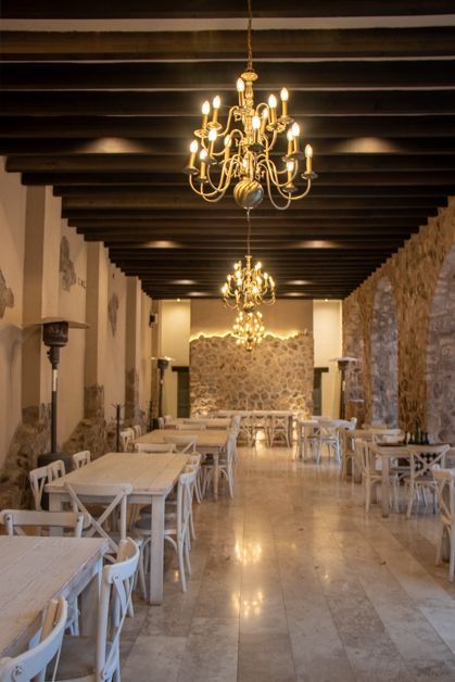 Elegant dining room with chandeliers, white tables, stone walls, and wooden ceiling beams.