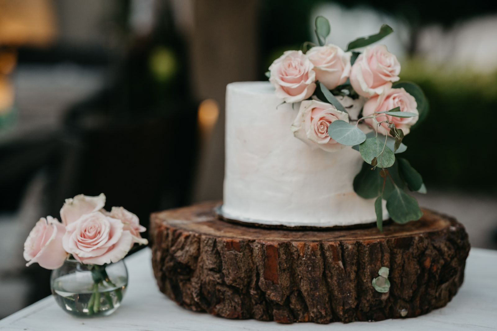 White cake decorated with pink roses on a wood slice cake stand. Small vase of roses in the foreground.