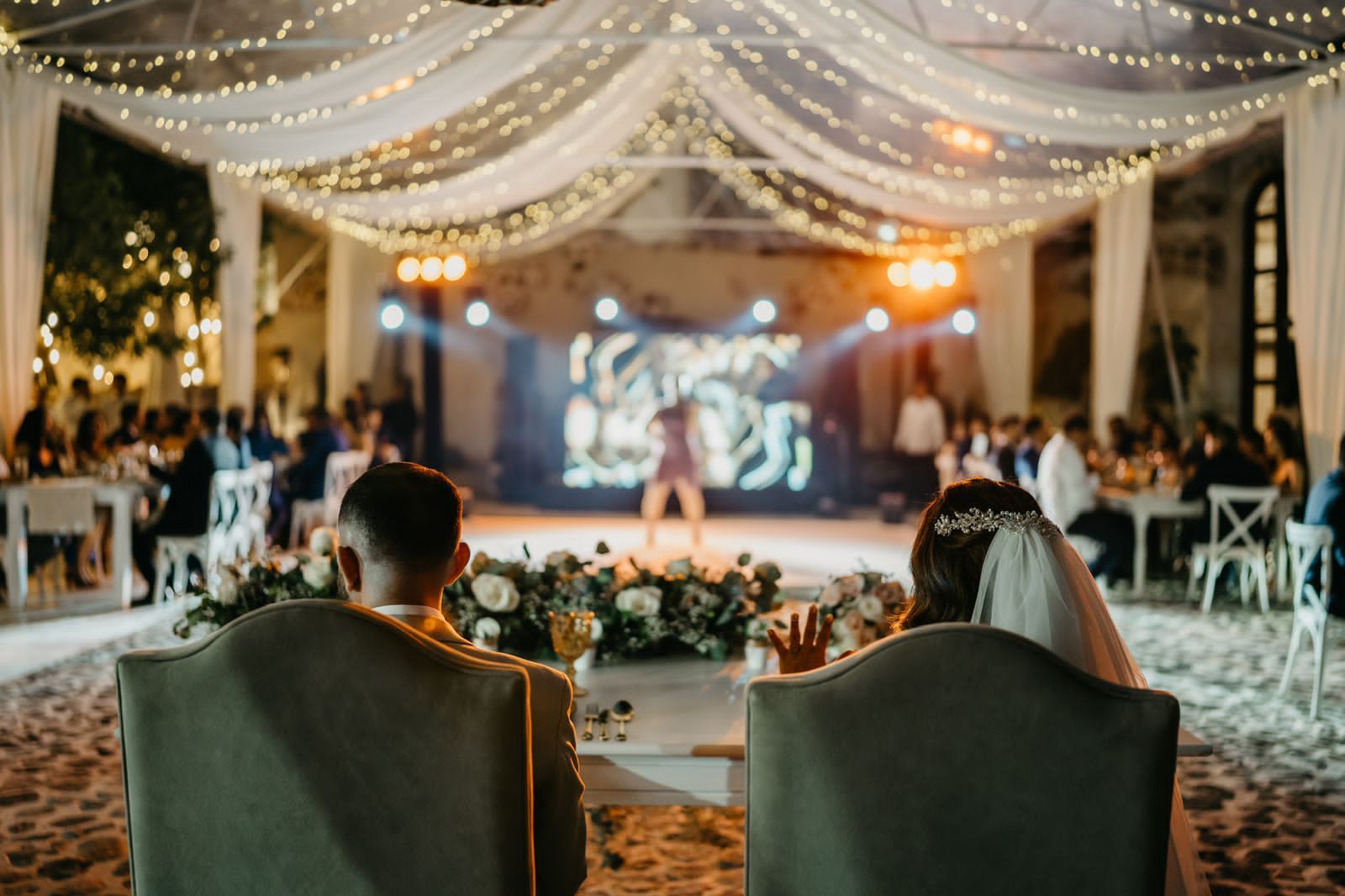 Bride and groom at a wedding reception, facing a stage with a performer. Soft lighting, white draping.