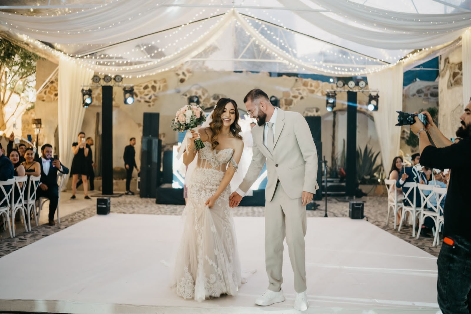 Newlyweds on a dance floor, she in a white dress, he in a light suit. Guests and decorations surround them.
