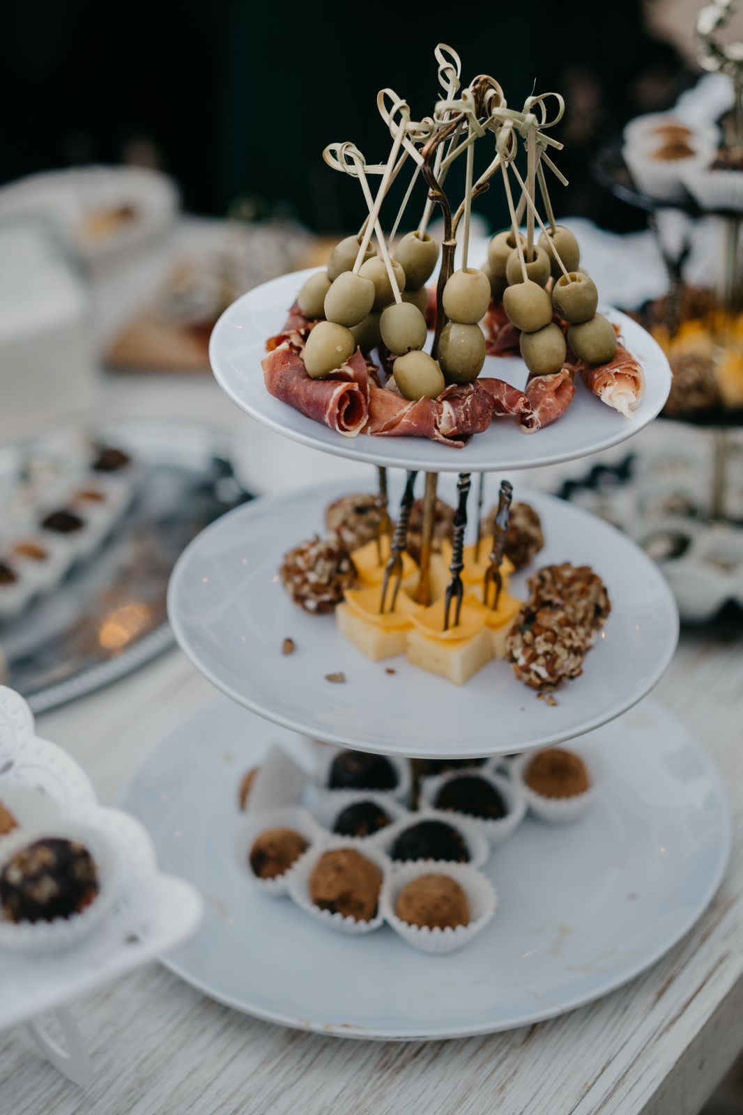 Three-tiered serving stand with olives and prosciutto skewers, pineapple bites, and chocolate treats.