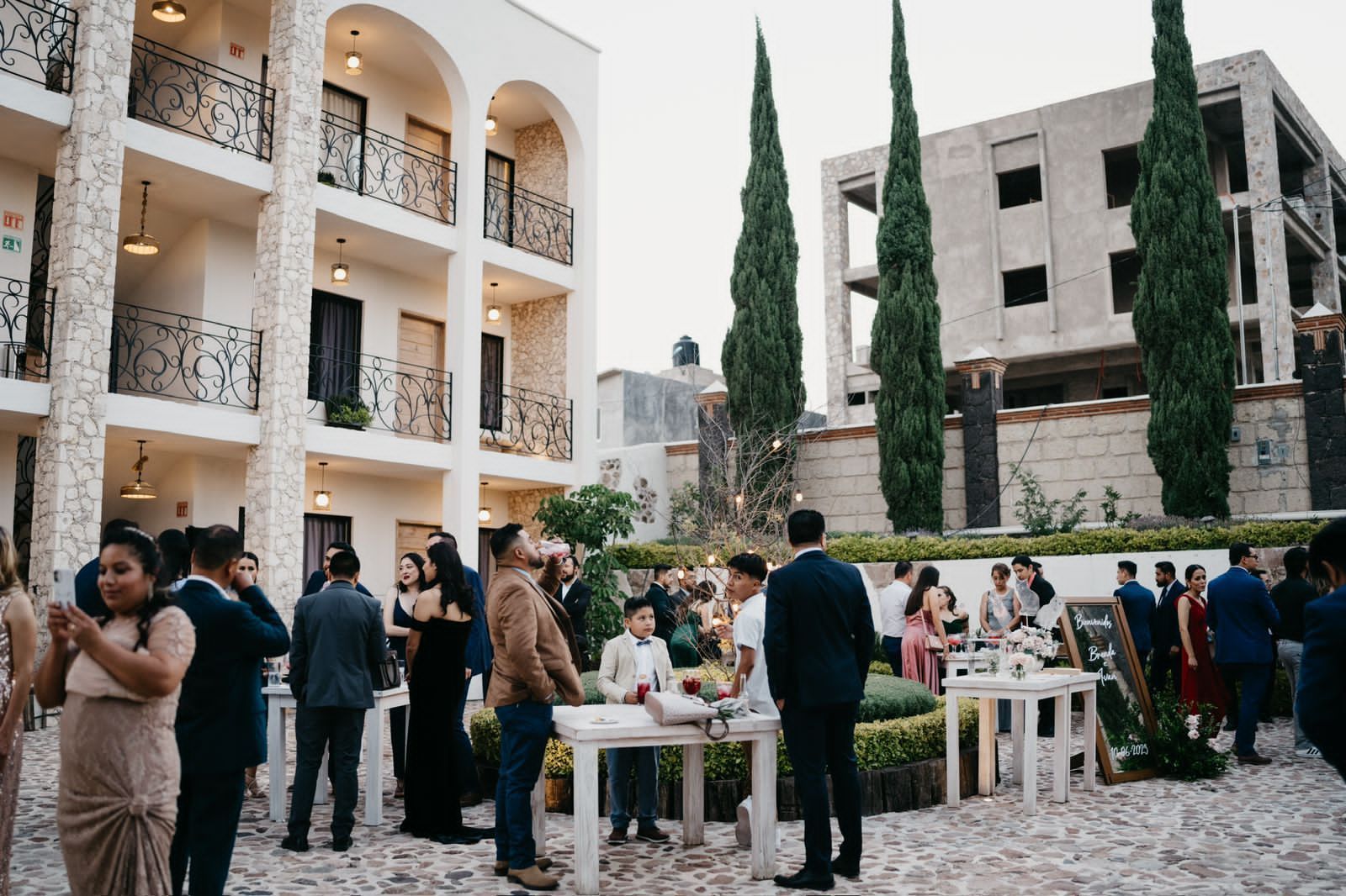 Guests at an outdoor event near a white building with arches and balconies, with tall trees and tables.