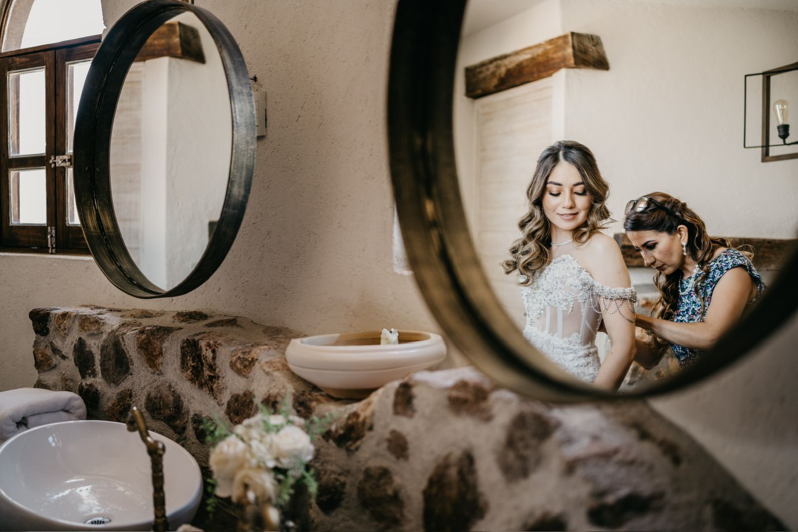 Bride in wedding dress, being helped by woman; reflected in a circular mirror.