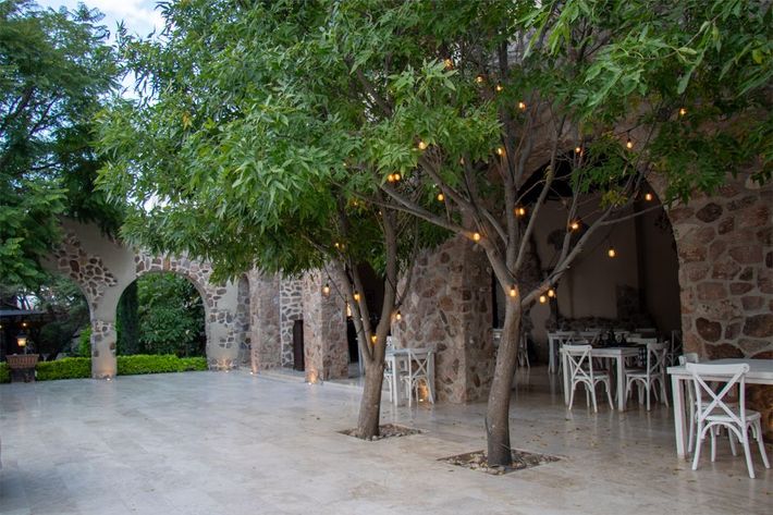 Outdoor patio with stone arches and trees lit with string lights; white tables and chairs.