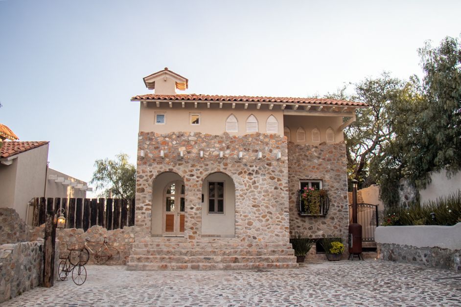 Stone house with arched entryway, steps, and small tower; beige walls, tile roof, cobblestone driveway.