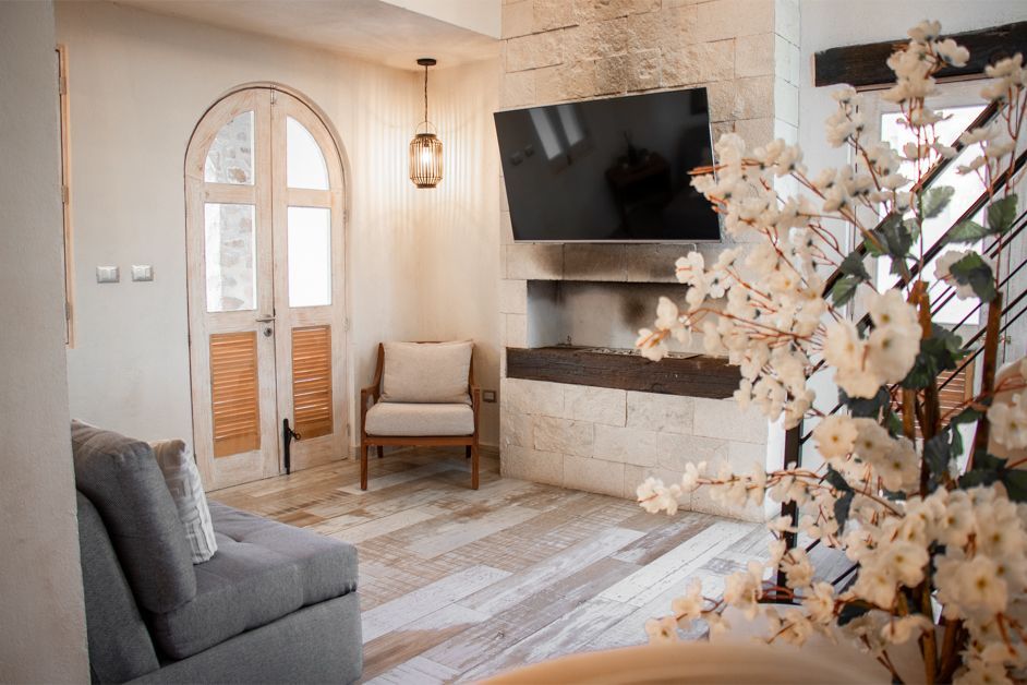 Cozy living room with gray sofa, chair, TV, and white flowering branches. Light wood flooring and arched doorway.