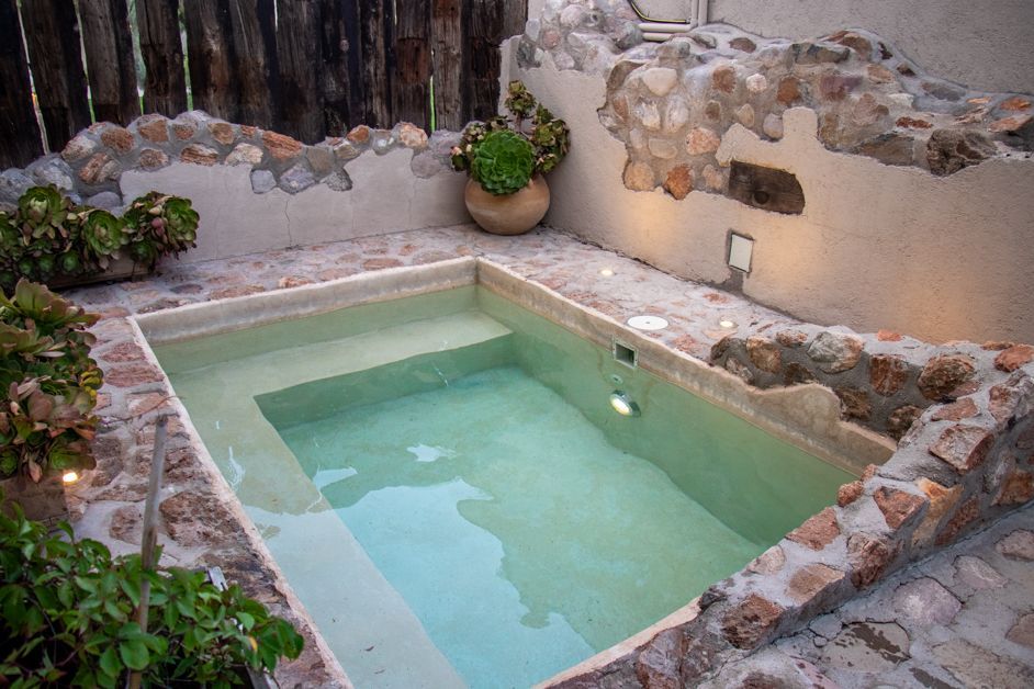 Small outdoor spa with light green water surrounded by stone walls, plants, and a wooden fence.