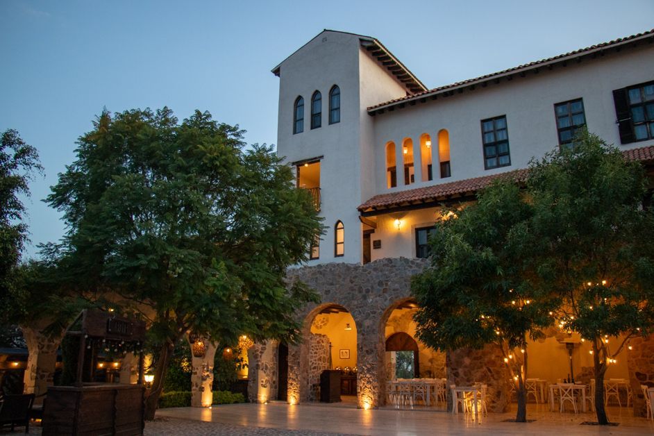 White building with arched stone entrance at dusk, trees with lights, outdoor seating.
