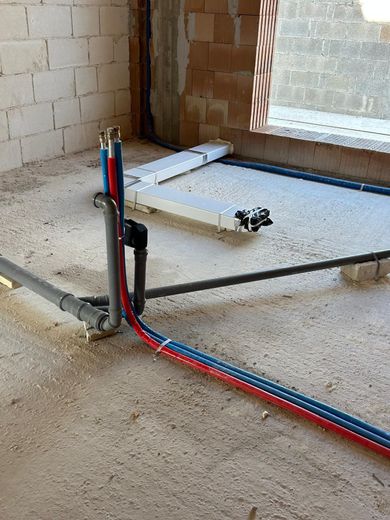 Pavimento di un cantiere edile con tubi rossi e blu a vista, una trincea e piastrelle impilate vicino a una finestra.