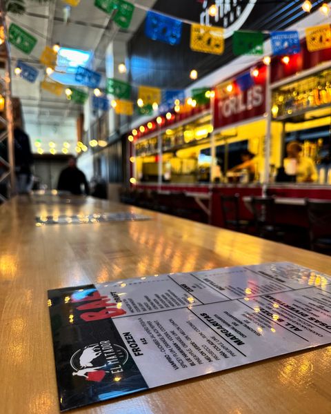 Menu on a wooden table in a colorful Mexican restaurant with paper flags and bright lights.