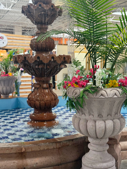 A tiered brown fountain with splashing water, surrounded by flower-filled stone planters in an indoor setting.