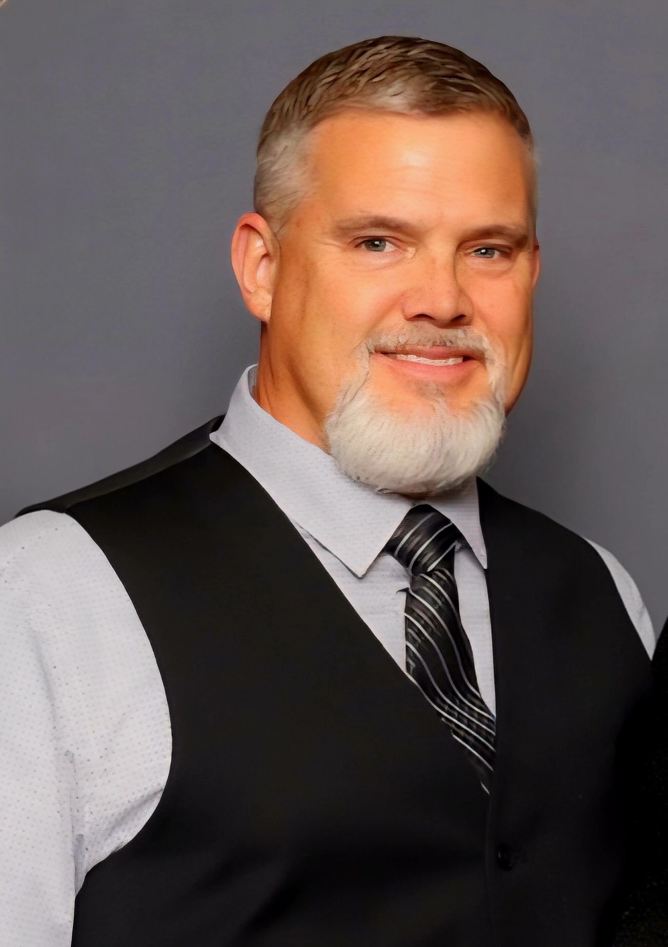 Man with graying hair and beard, wearing a vest and tie, smiling.