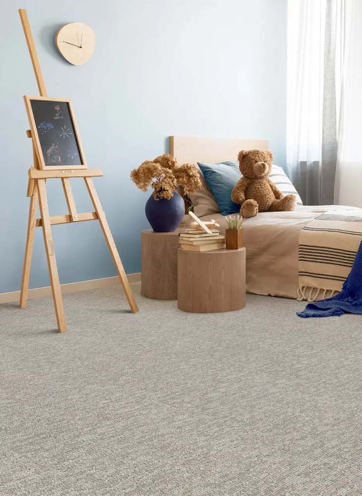 Bedroom with light blue walls, wooden furniture, and gray speckled carpet. A bed with a teddy bear, easel, and round side tables.
