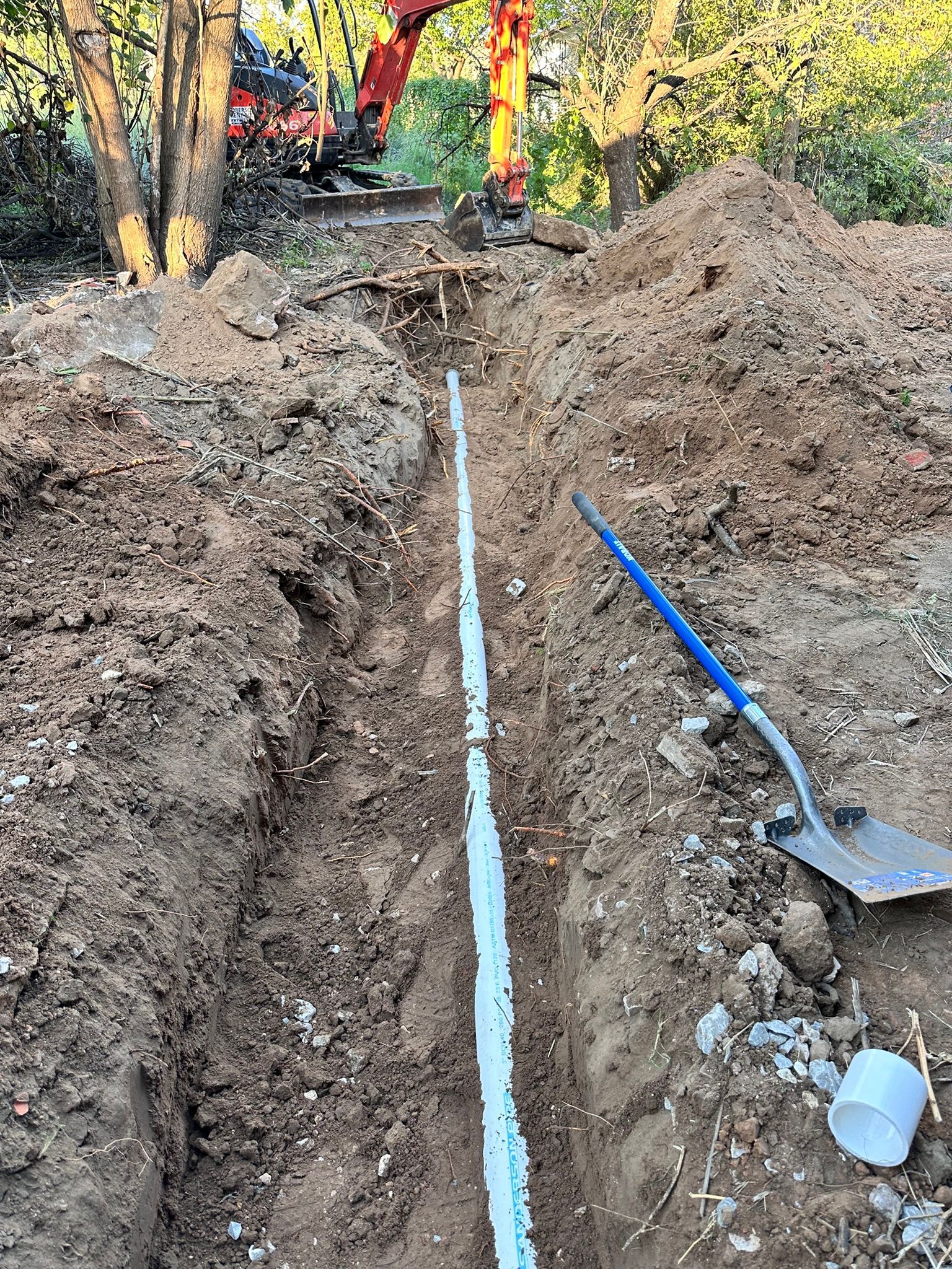 A trench dug in the ground with white pipe inside, shovel, dirt pile, and excavator in the background.