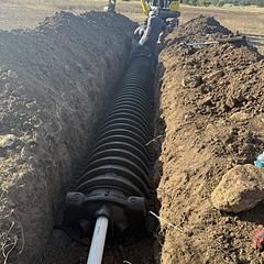 Workers installing corrugated black pipe in a deep trench dug in a field.