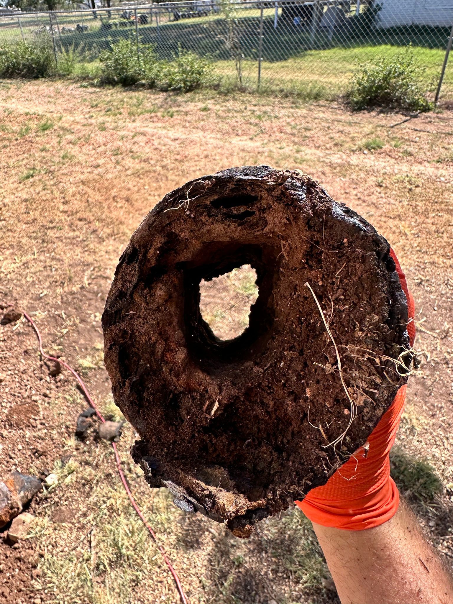 Hand holding a dark, hollowed-out wooden object with a square hole. Outdoors on brown ground.