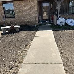 Concrete walkway leading to a brick house entrance. Brown dirt surrounds the path.