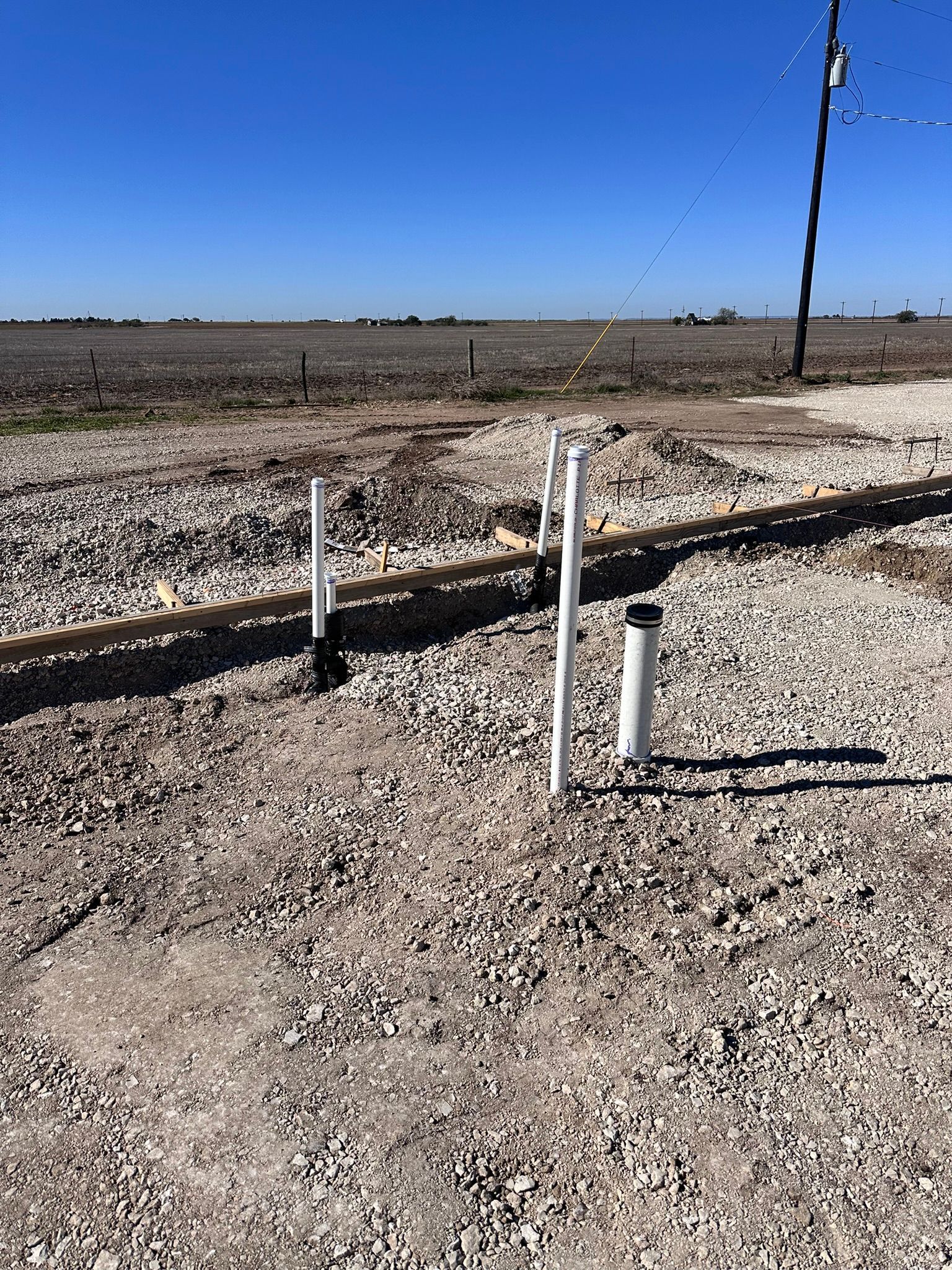 White PVC pipes protruding from gravel ground, likely for plumbing, in an outdoor rural setting.