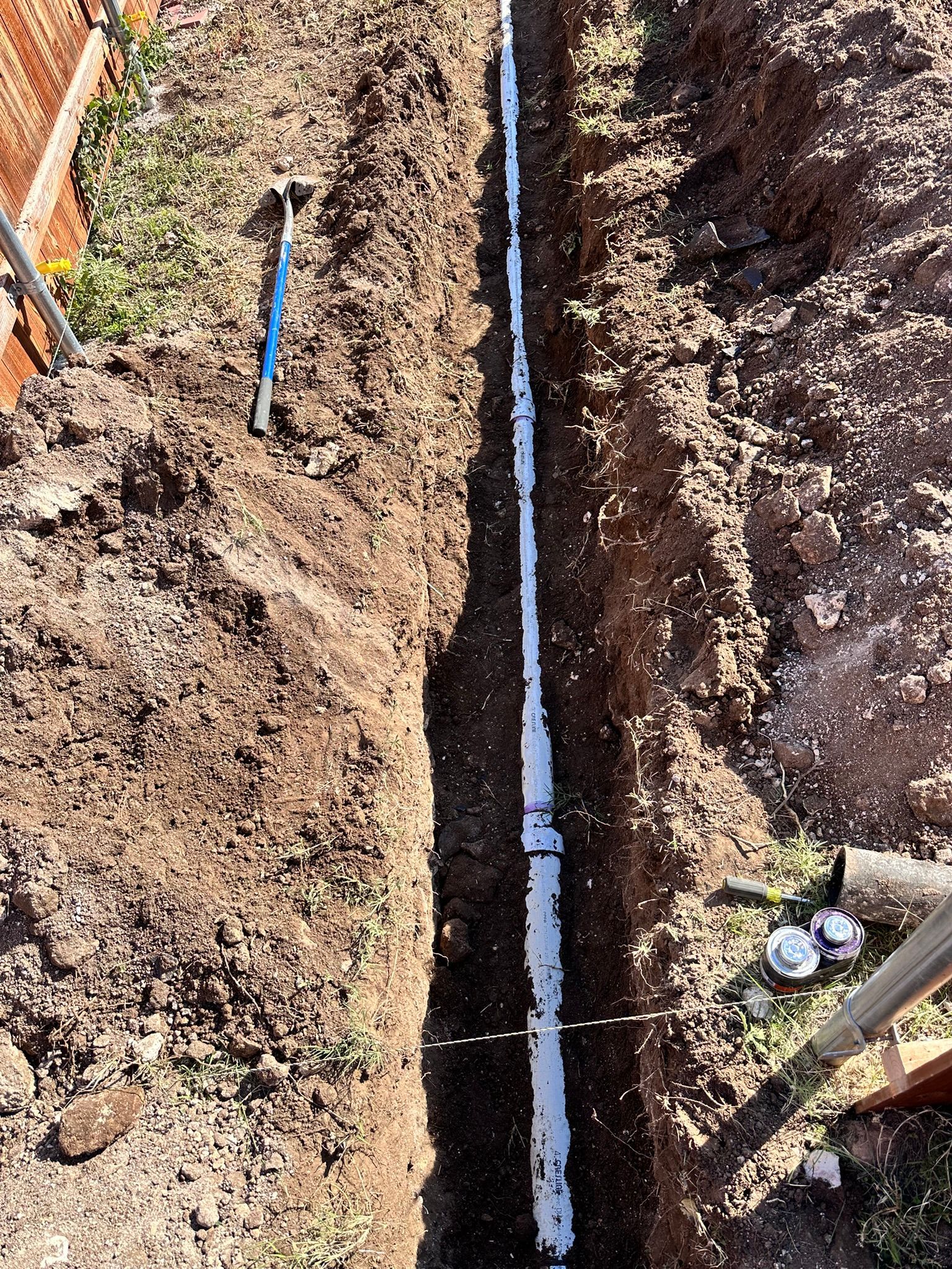 Trench with white PVC pipes laid out, along a fence in a brown yard.
