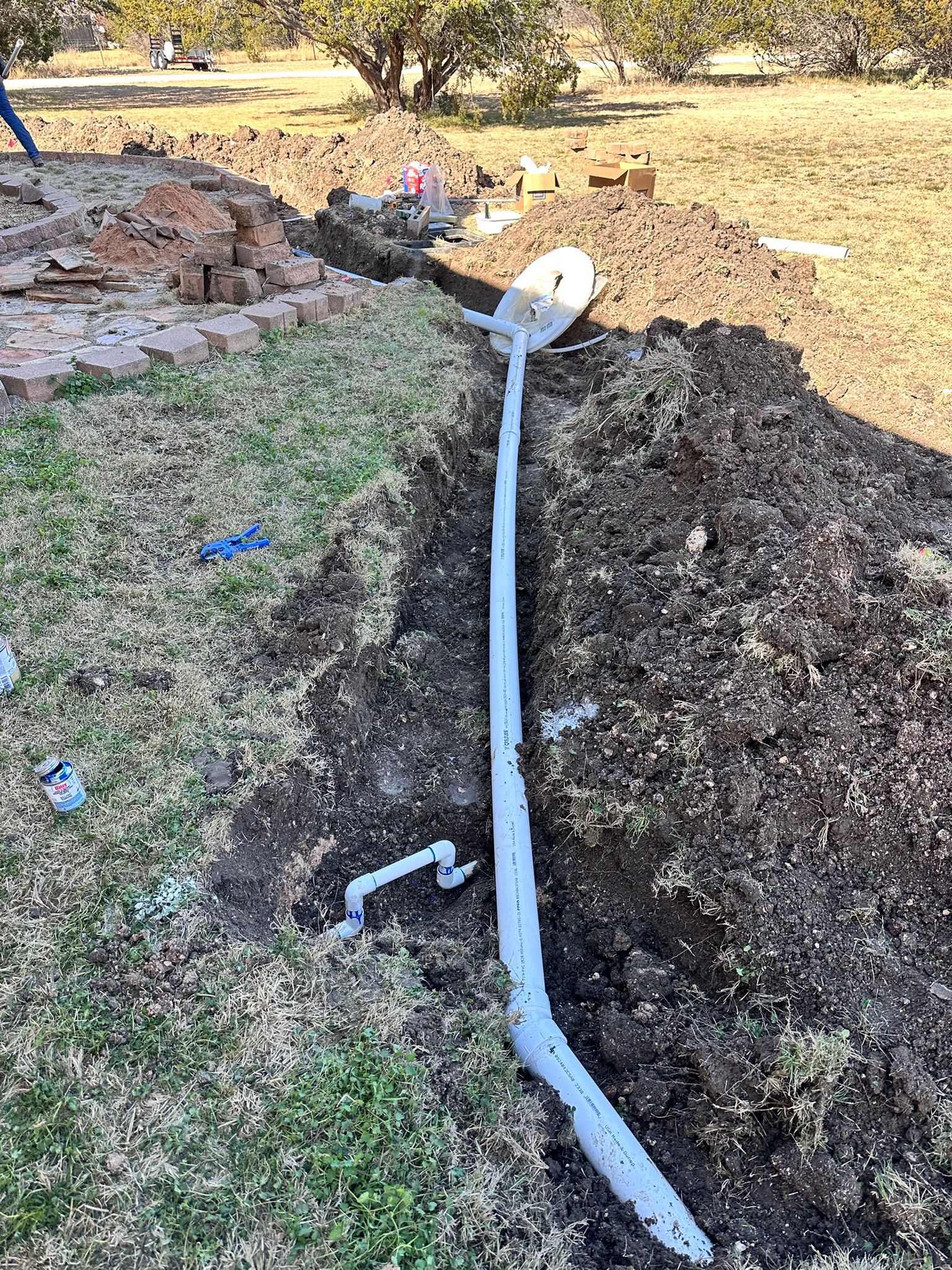 Trench dug in a yard with exposed gray pipes. Bricks and dirt pile visible.
