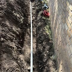 Trench dug next to a brick wall, with a metal measuring stick inside.