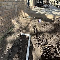 A construction site shows a trench with white pipes near a brick wall and concrete patio.
