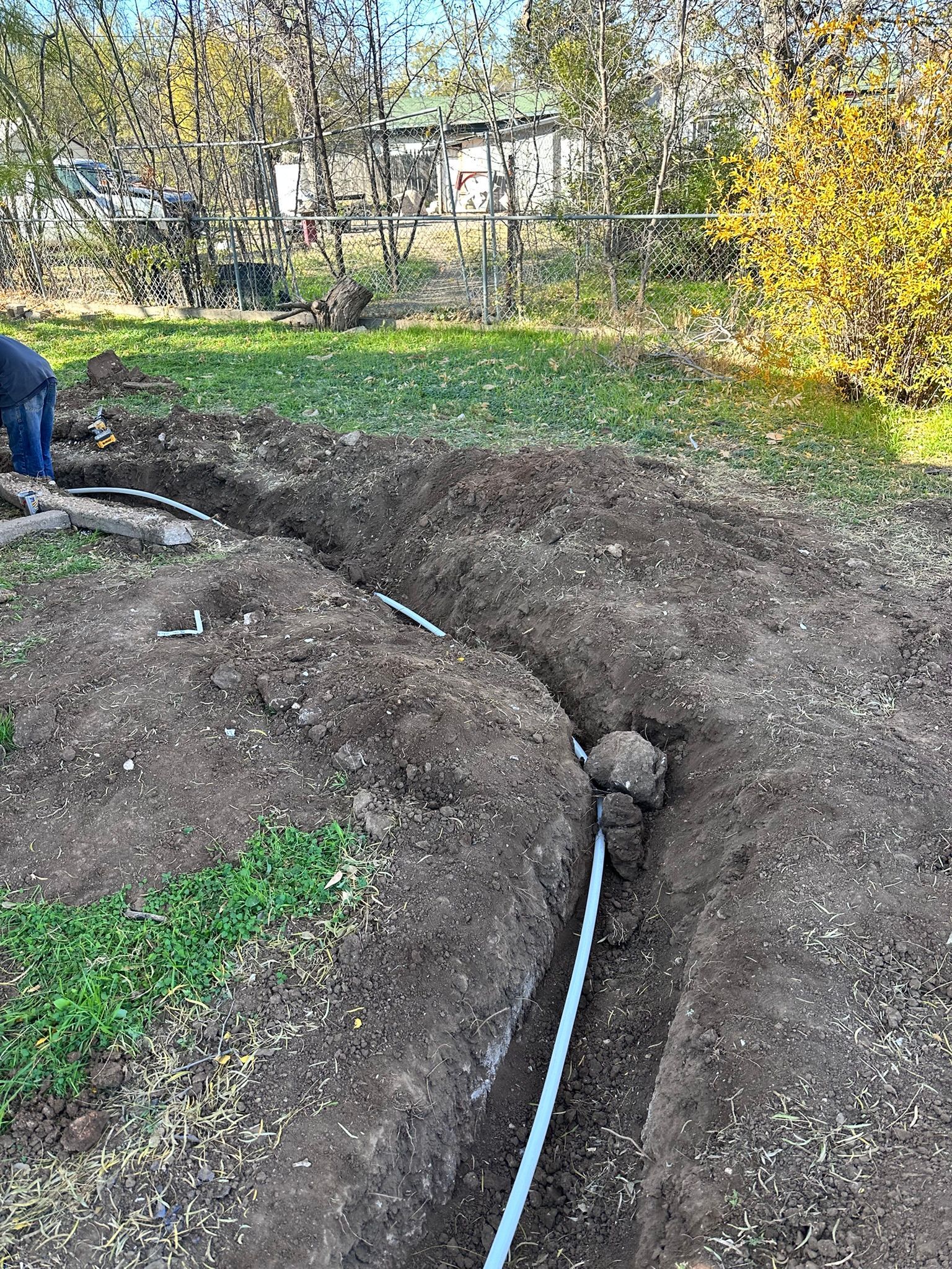 Person digging a trench in a grassy yard, installing a blue pipe; trees and a house in the background.