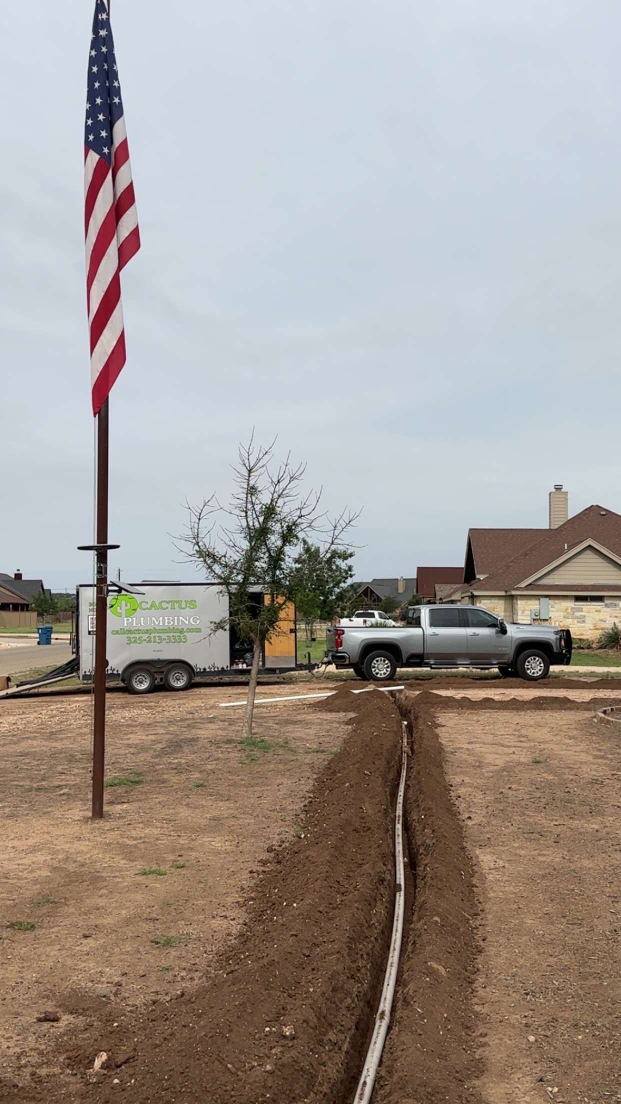 A silver truck pulling a trailer, trench with a cable, American flag, and a house.