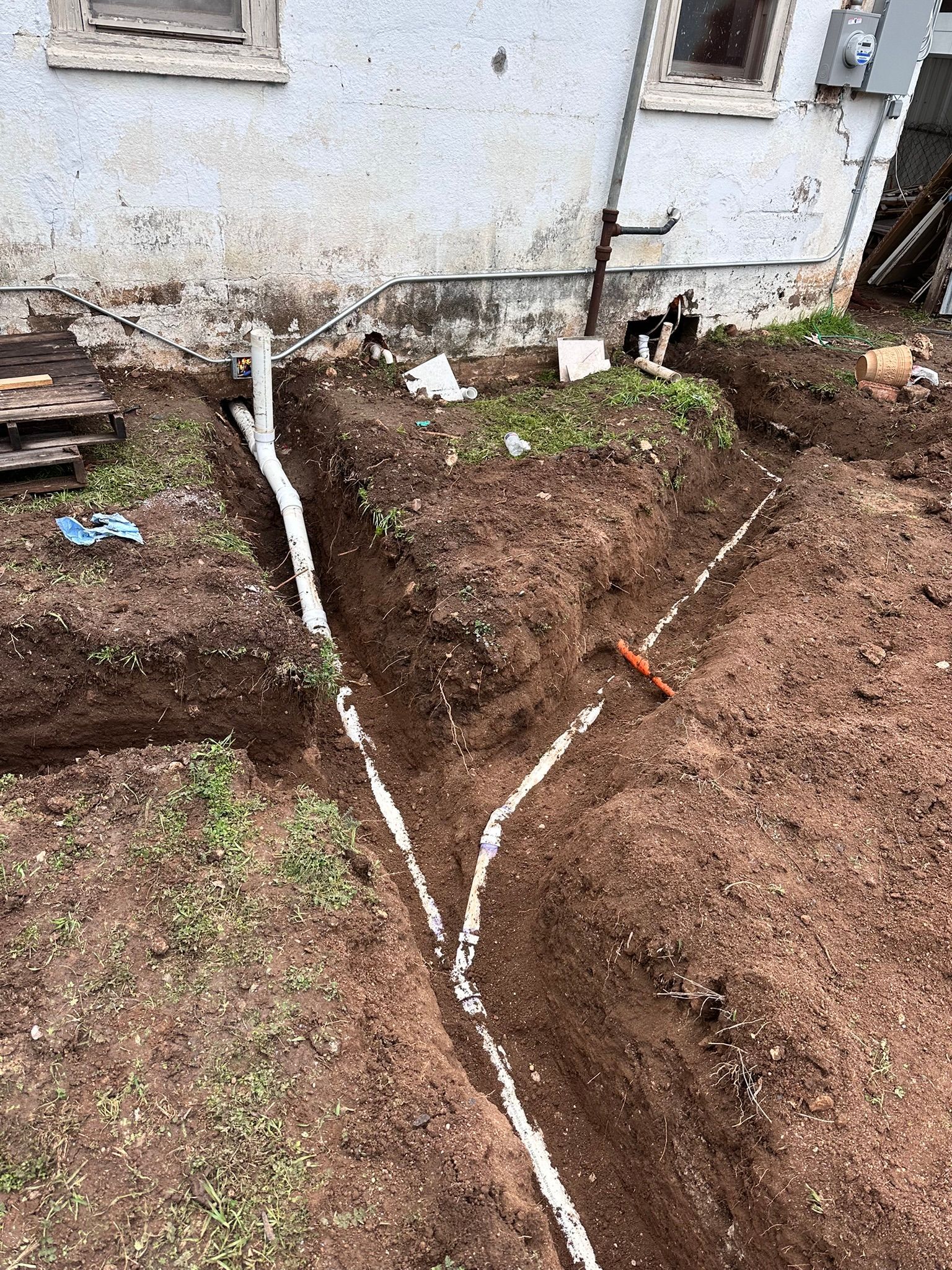 Excavated trenches with white pipes, likely for drainage, next to a building. Earth and grass are visible.