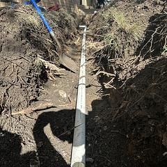 Trench with a white pipe running through it, dug in dirt. A blue shovel rests in the dirt to the left.