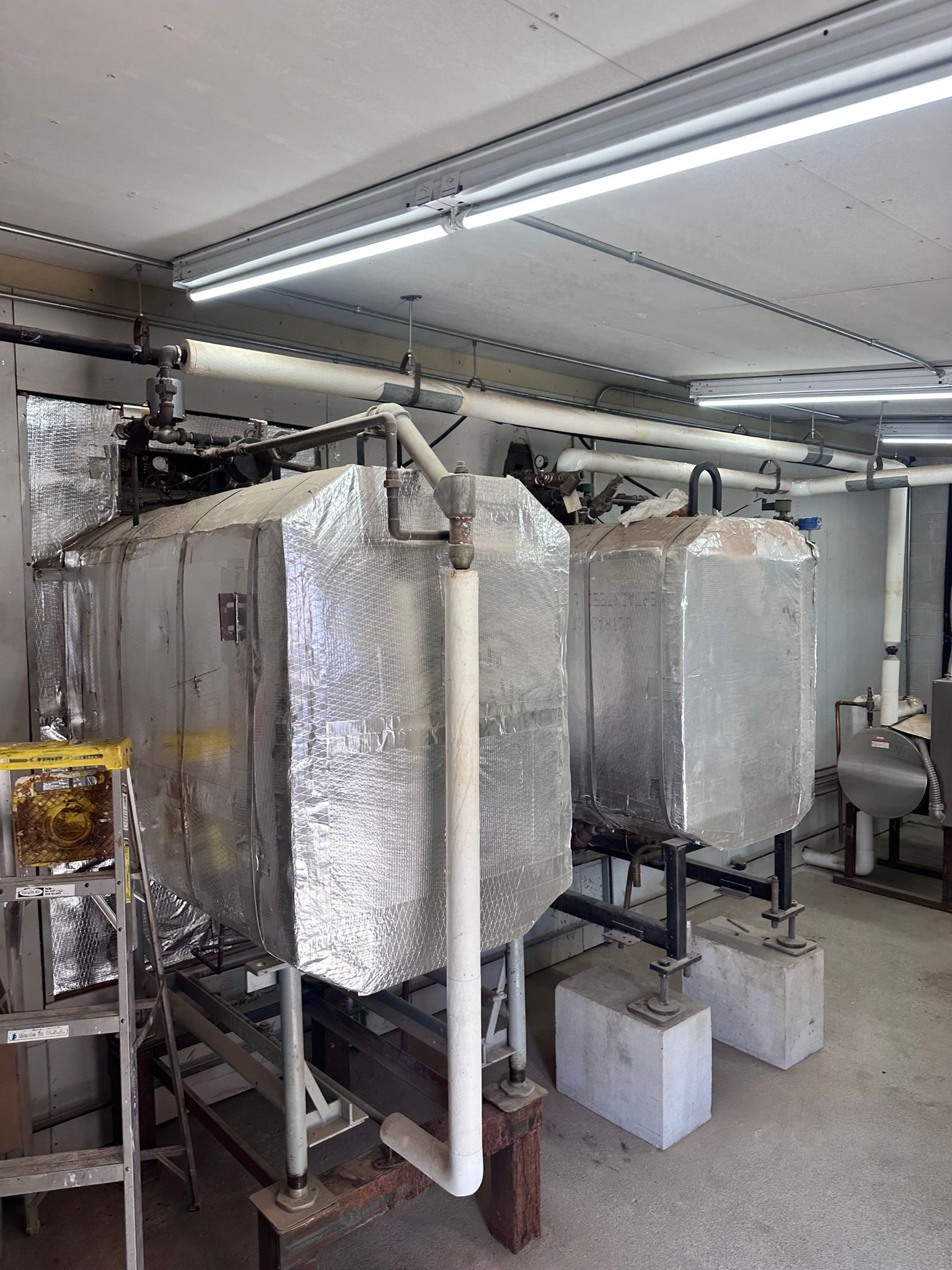 Two insulated industrial tanks in a room with pipes. Silver insulation, white pipes, and concrete blocks.