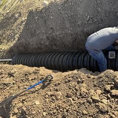 A person installing corrugated drainage pipe in an excavated trench. Brown dirt surrounds them.