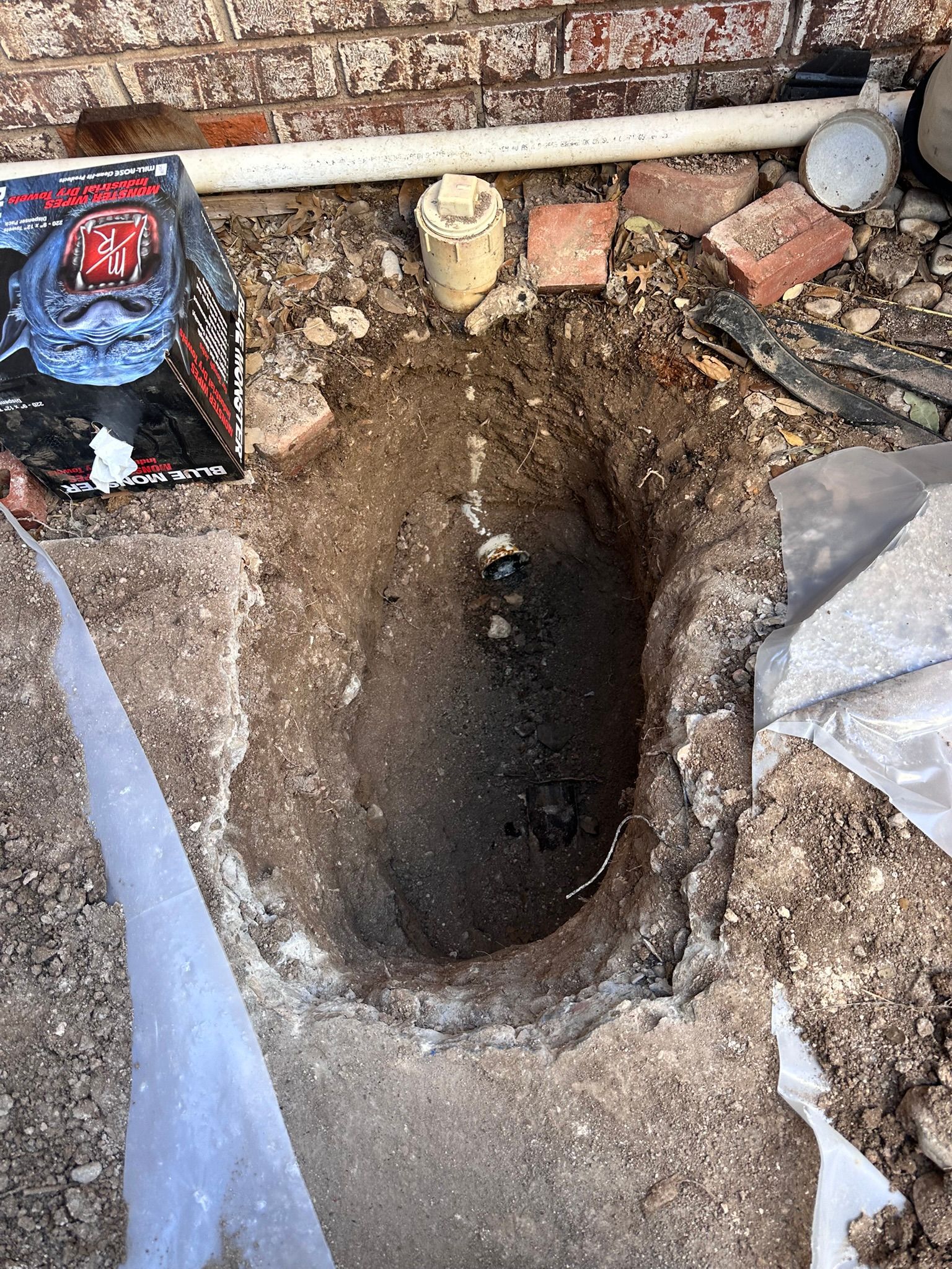 Hole dug in the ground, exposing a drain pipe. Red brick wall in the background.