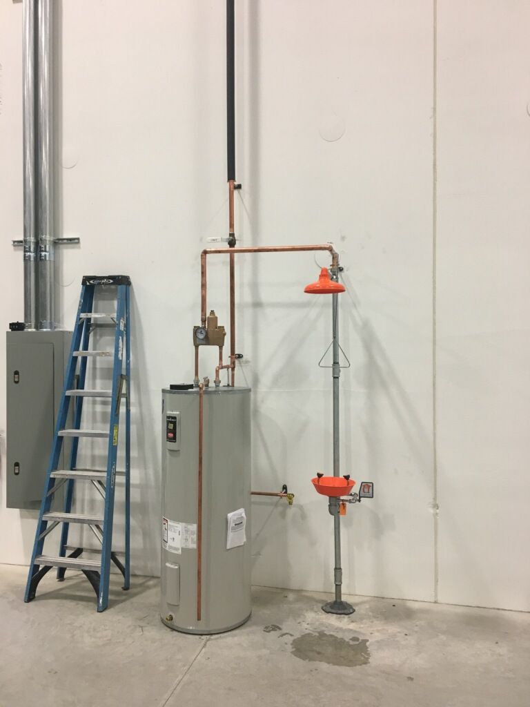 Water heater with safety shower and eyewash station next to a ladder and electrical panel in a room.