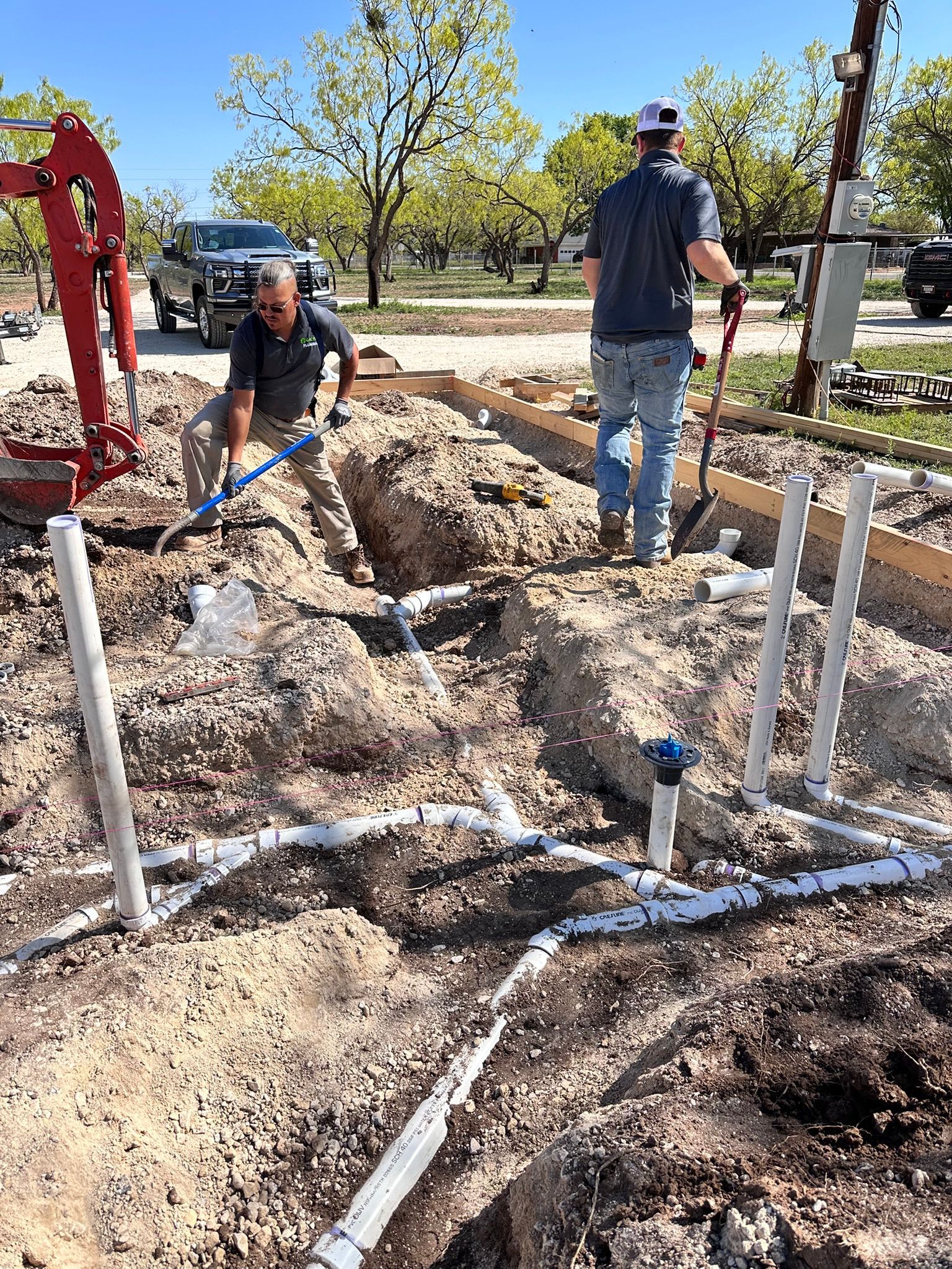 Two men installing plumbing pipes in a dirt trench on a sunny day.