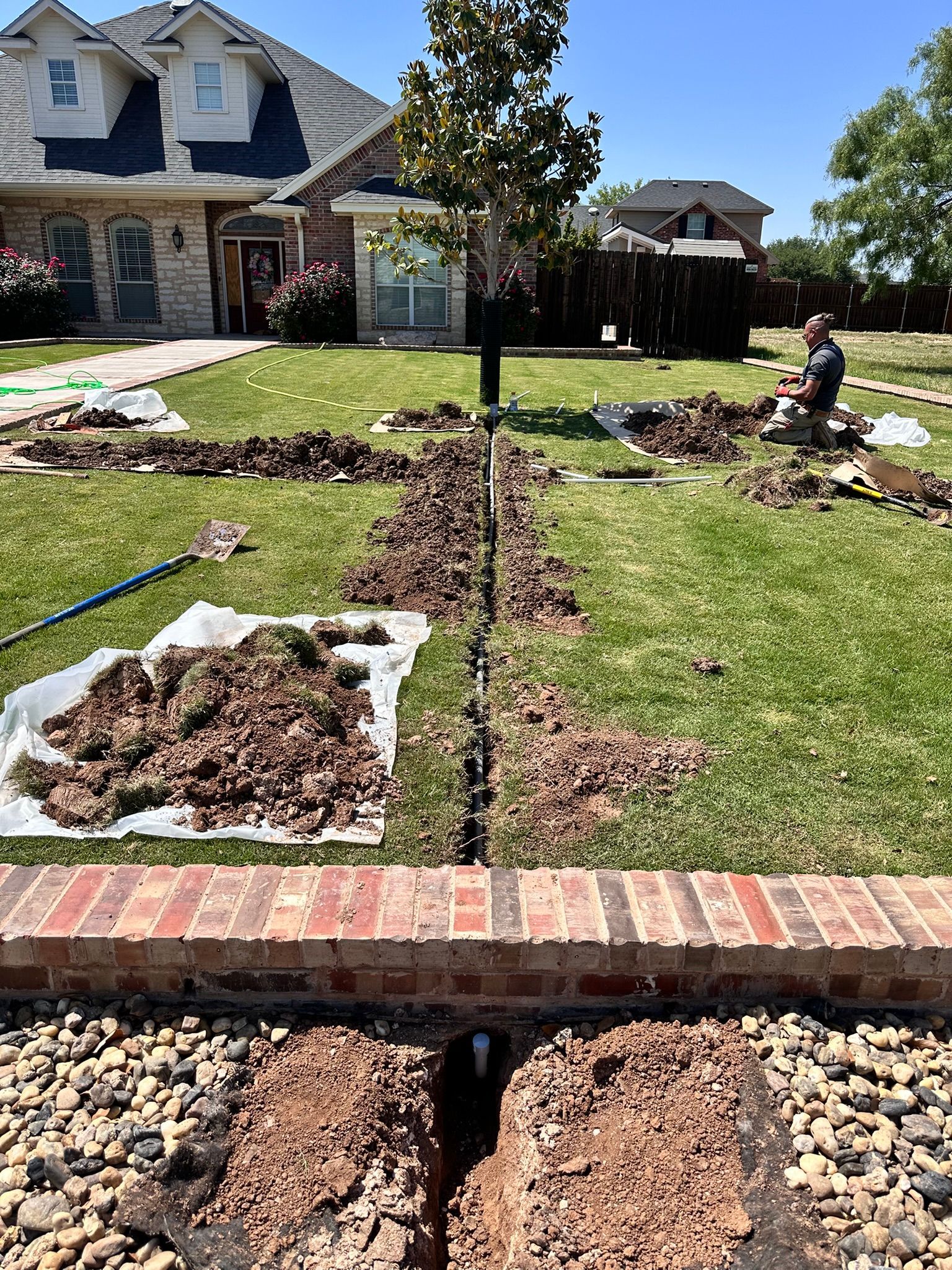 Lawn under construction with trenches, dirt, and a person working on a sprinkler system in front of a house.